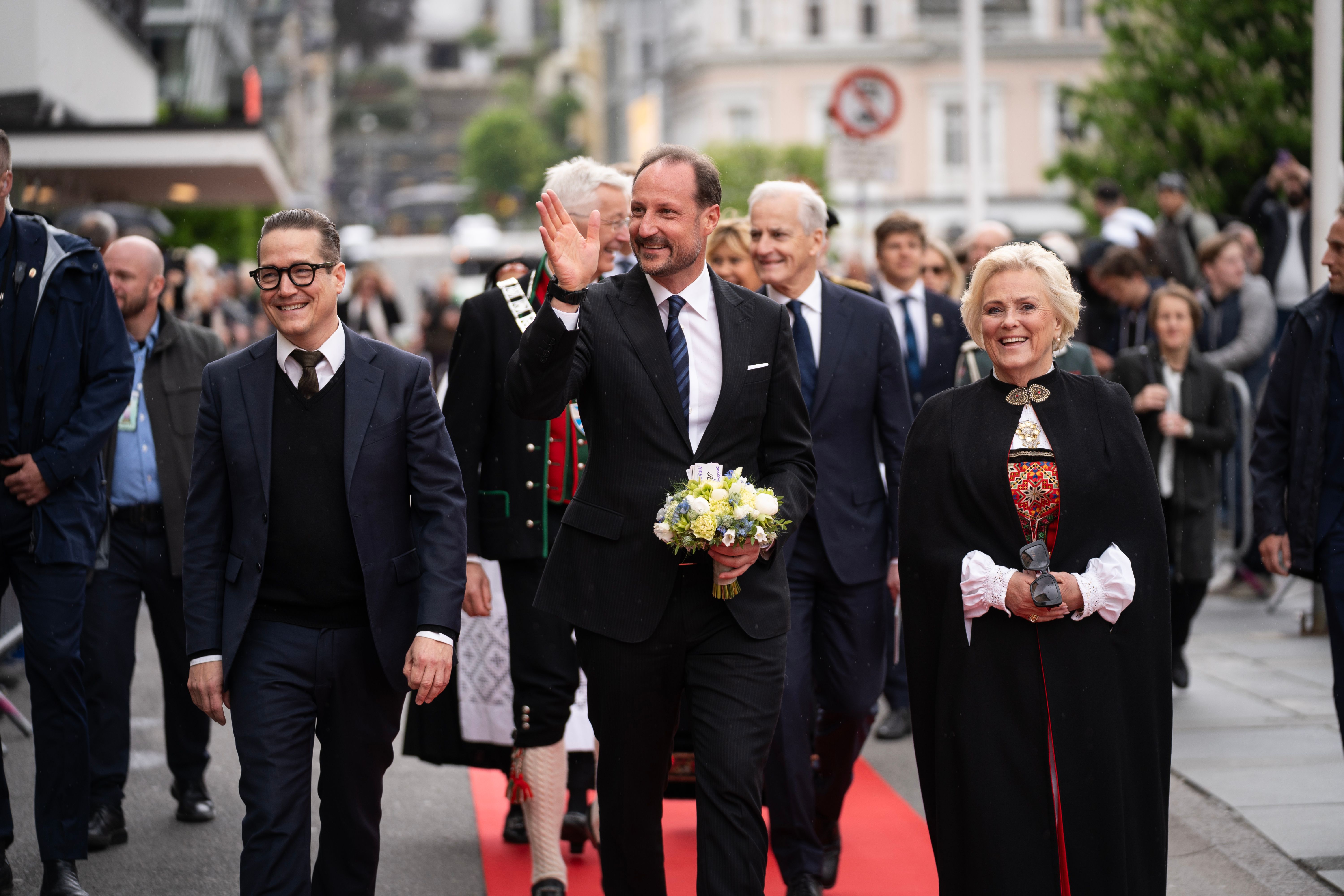 Kronprinsen kommer gående langs rød løper med blomster i hendene og mennesker rundt seg.