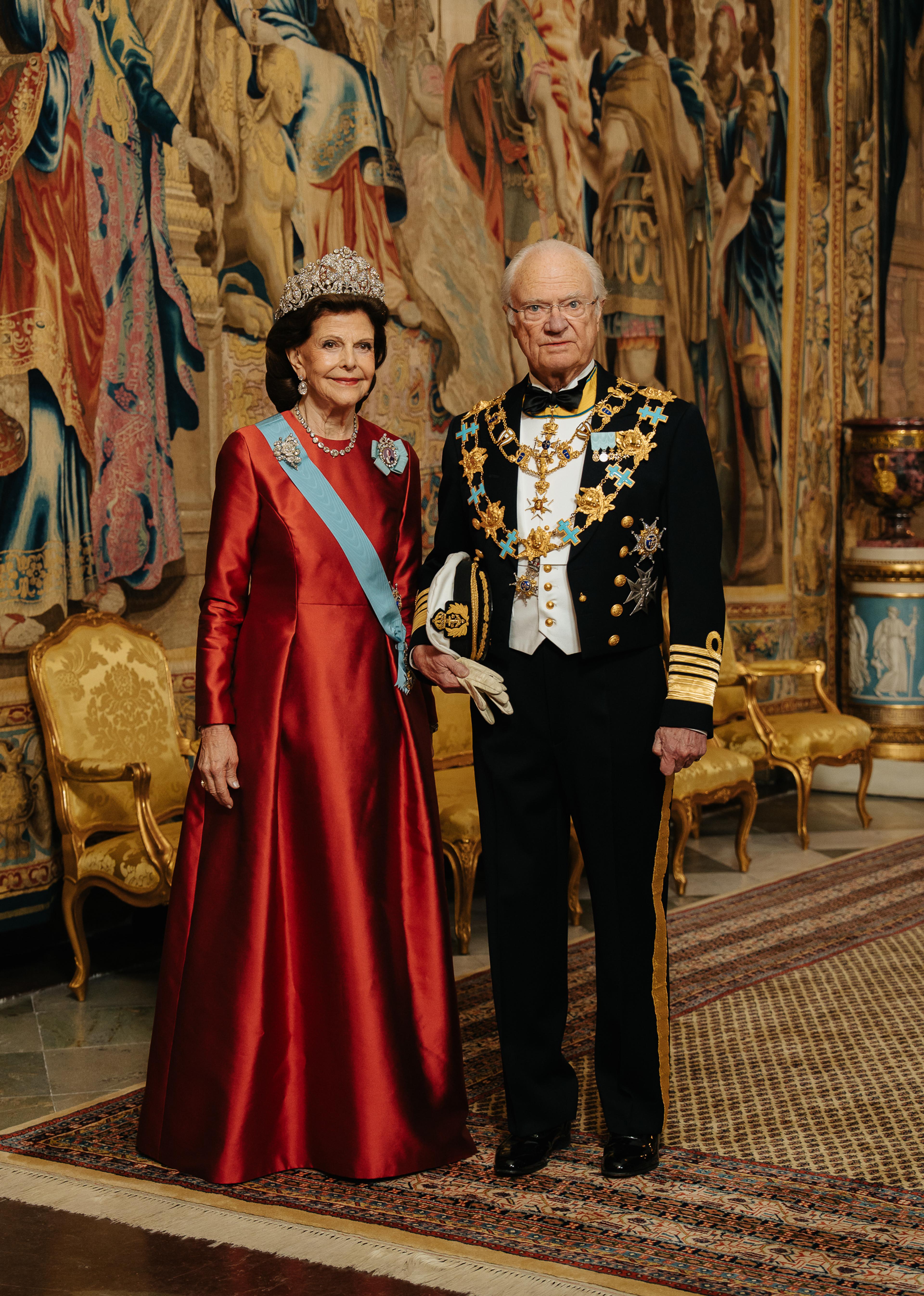 Kong Carl Gustaf og Dronning Silvia står i galla foran et stort veggteppe (gobelin). Dronningen er kledd i en fotsid, rød kjoe og Kongen i marinens gallauniform med ordenskjeder.