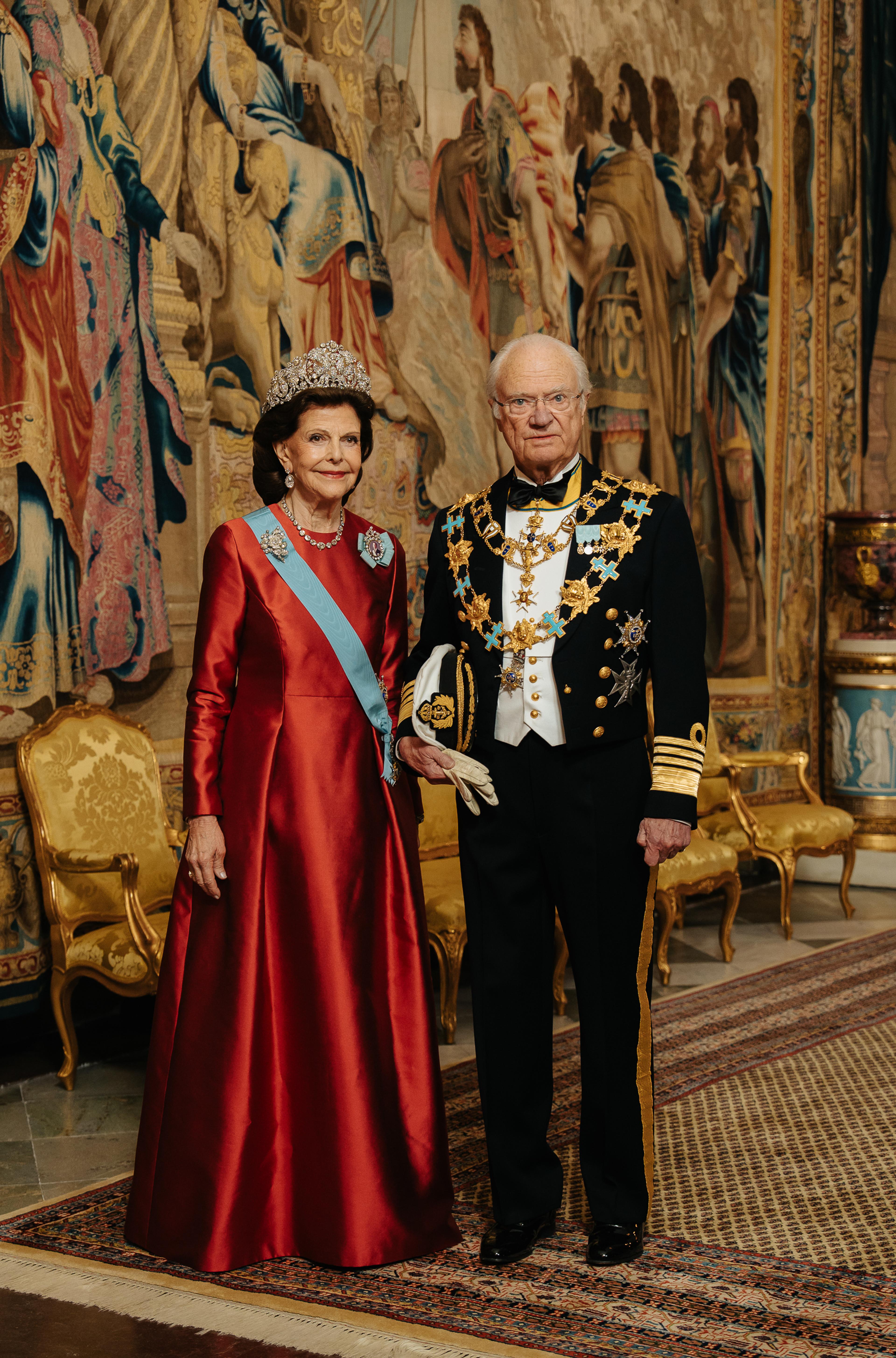 Kong Carl Gustaf og Dronning Silvia står i galla foran et stort veggteppe (gobelin). Dronningen er kledd i en fotsid, rød kjoe og Kongen i marinens gallauniform med ordenskjeder.