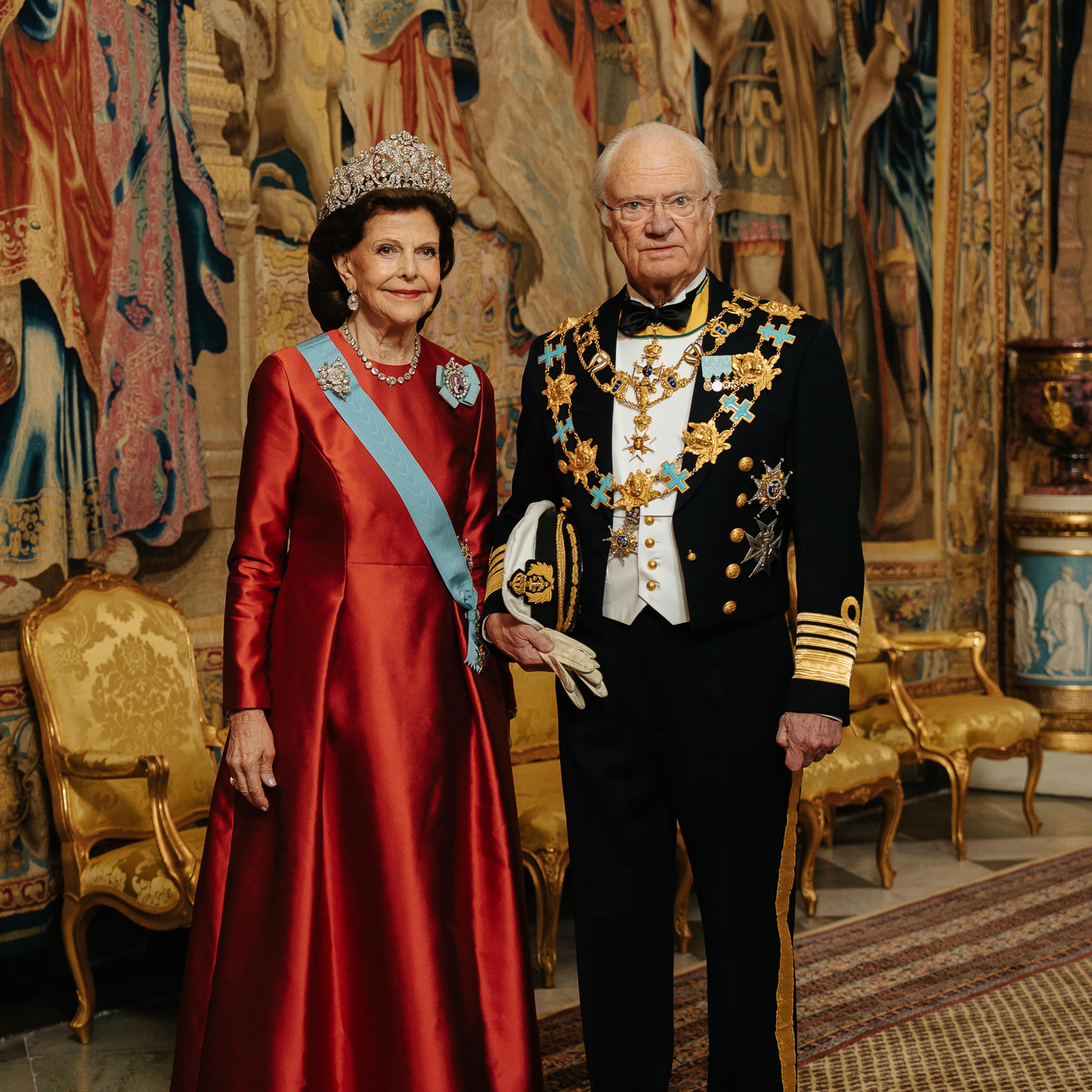 Kong Carl Gustaf og Dronning Silvia står i galla foran et stort veggteppe (gobelin). Dronningen er kledd i en fotsid, rød kjoe og Kongen i marinens gallauniform med ordenskjeder.