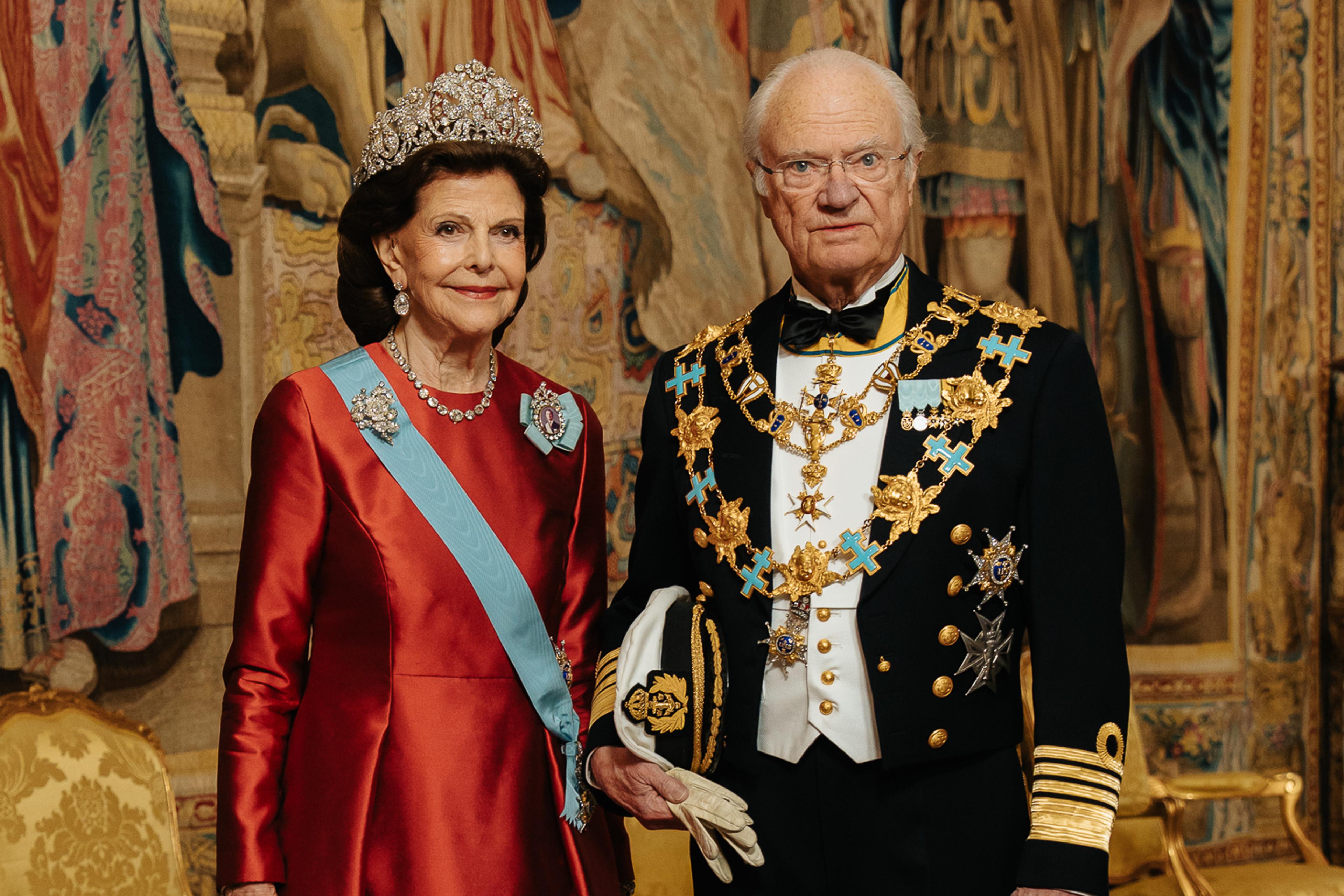 Kong Carl Gustaf og Dronning Silvia står i galla foran et stort veggteppe (gobelin). Dronningen er kledd i en fotsid, rød kjoe og Kongen i marinens gallauniform med ordenskjeder.