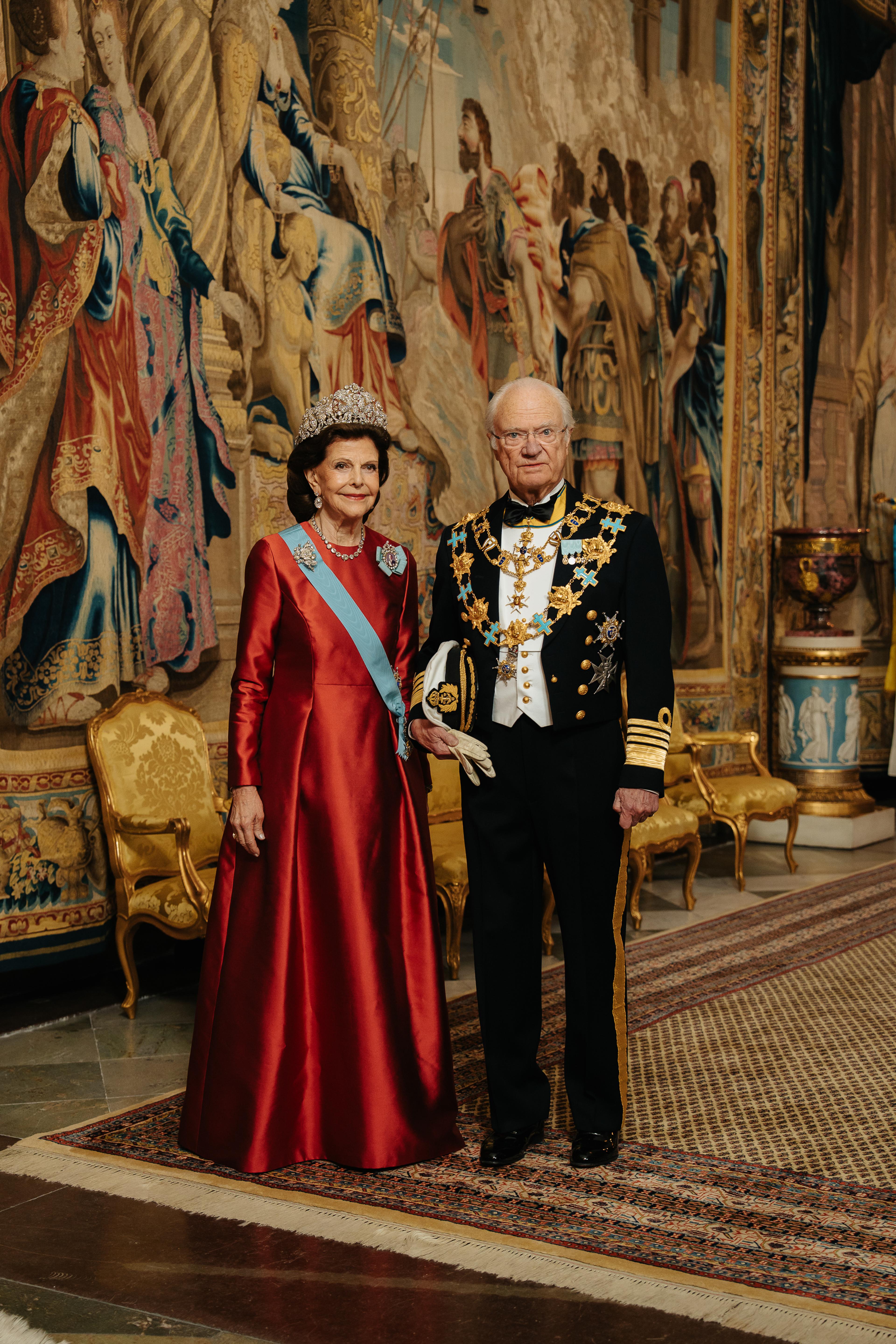 Kong Carl Gustaf og Dronning Silvia står i galla foran et stort veggteppe (gobelin). Dronningen er kledd i en fotsid, rød kjoe og Kongen i marinens gallauniform med ordenskjeder.