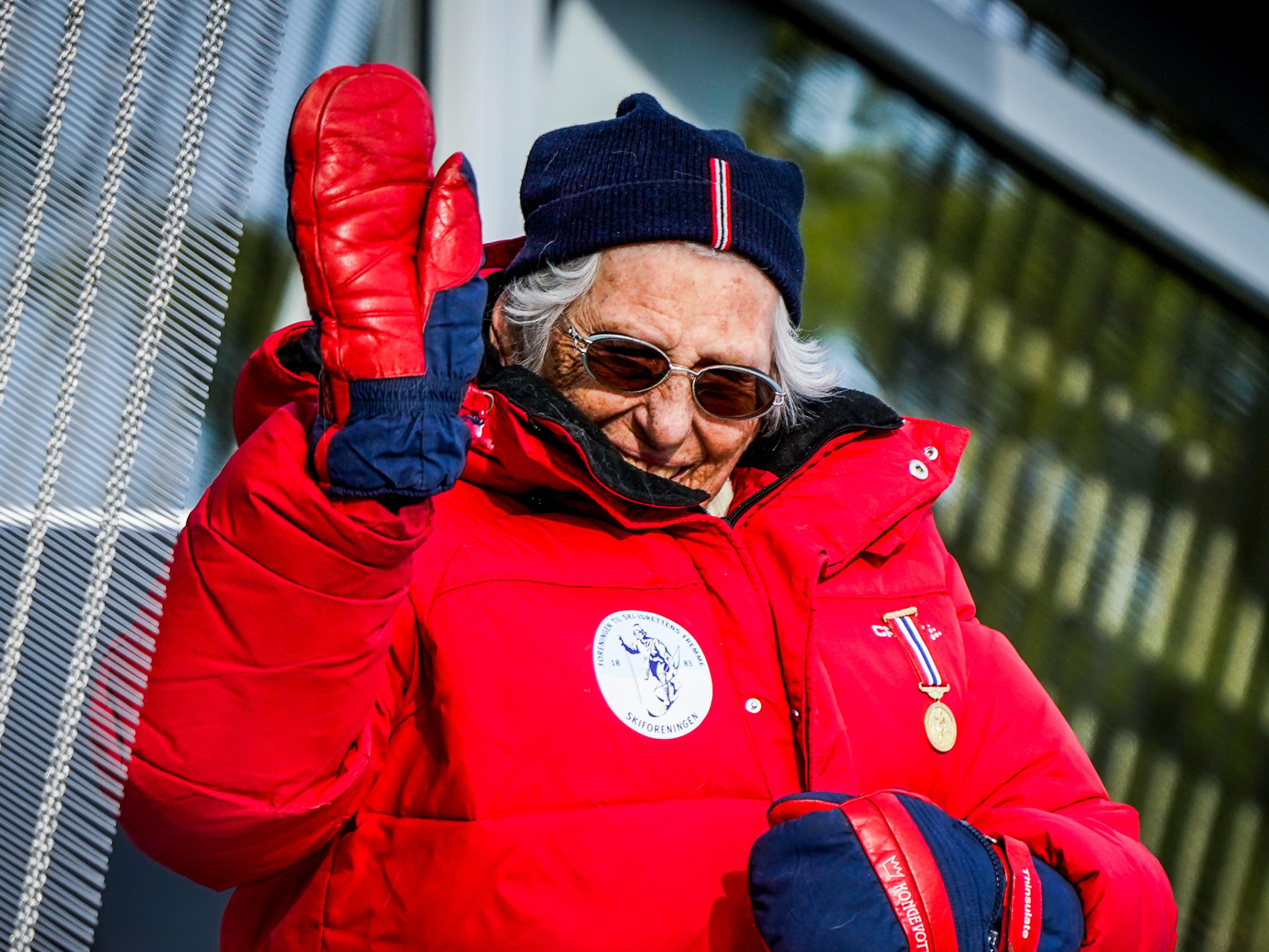 Prinsessen vinker fra tribunen, godt kledd med solbriller og varm, rød jakke.