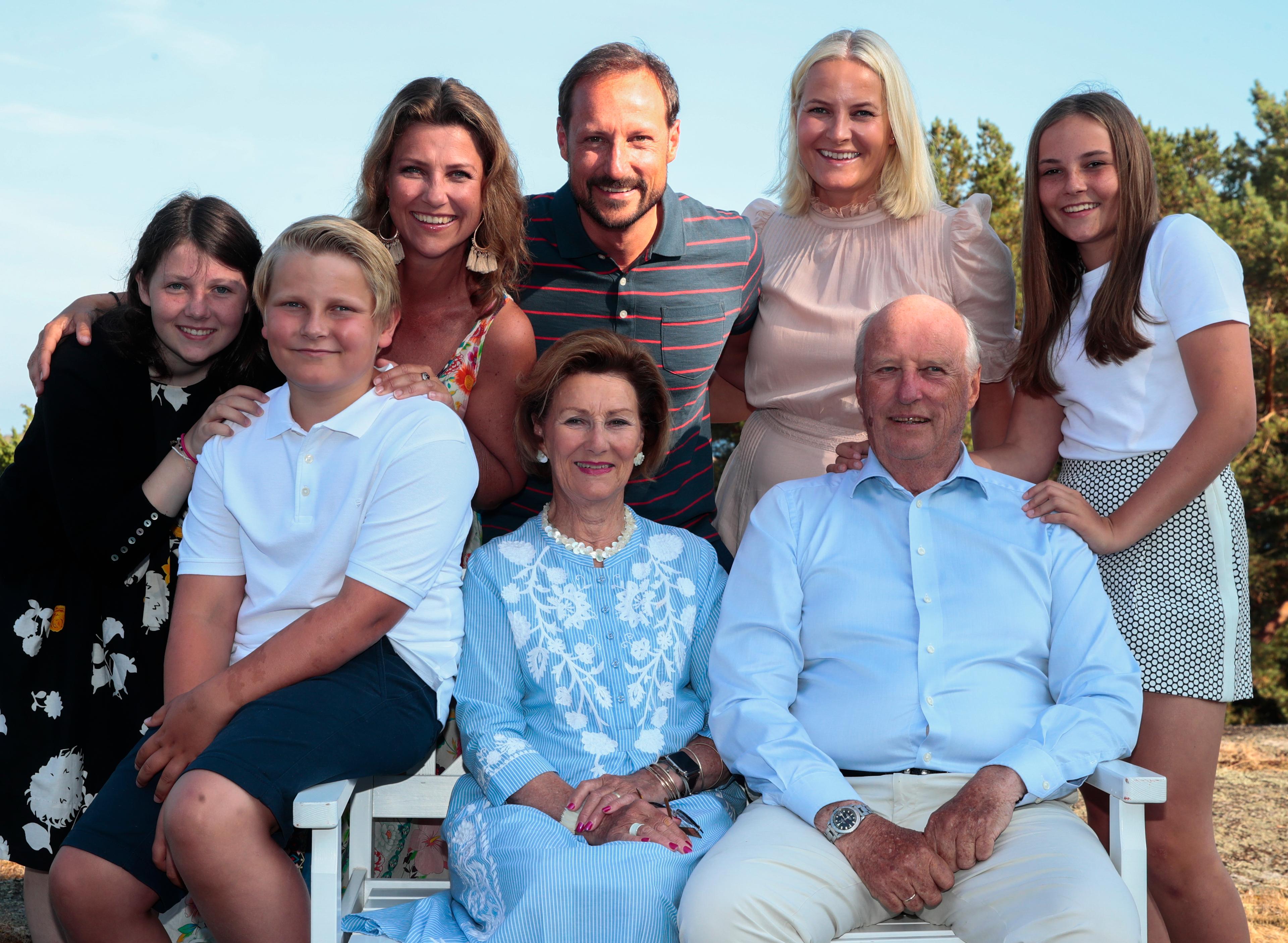Kong Harald og Dronning Sonja sitter på en hvit benk med barn, svigerdatter og barnebarn rundt seg.