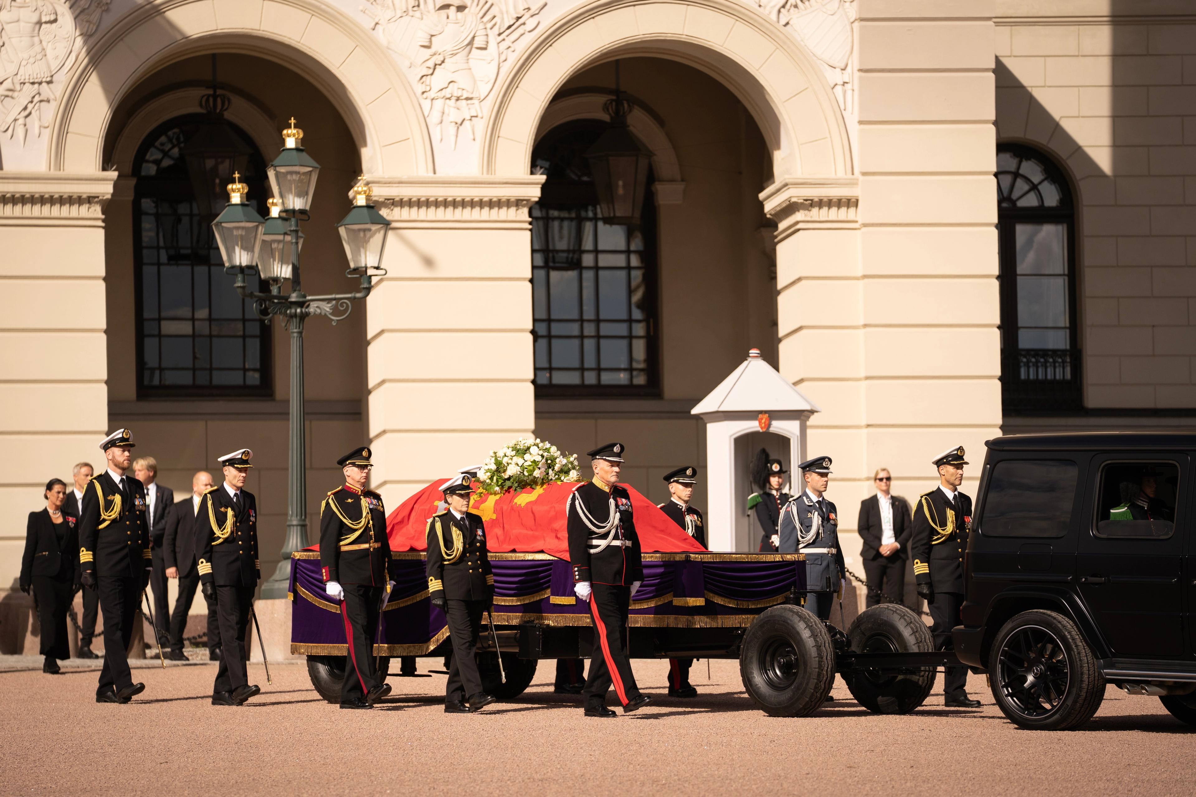 Kong Haralds båre med Kongeflagget drapert over ligger på katafalk. Kongens adjutantstab går på hver side av katafalken.