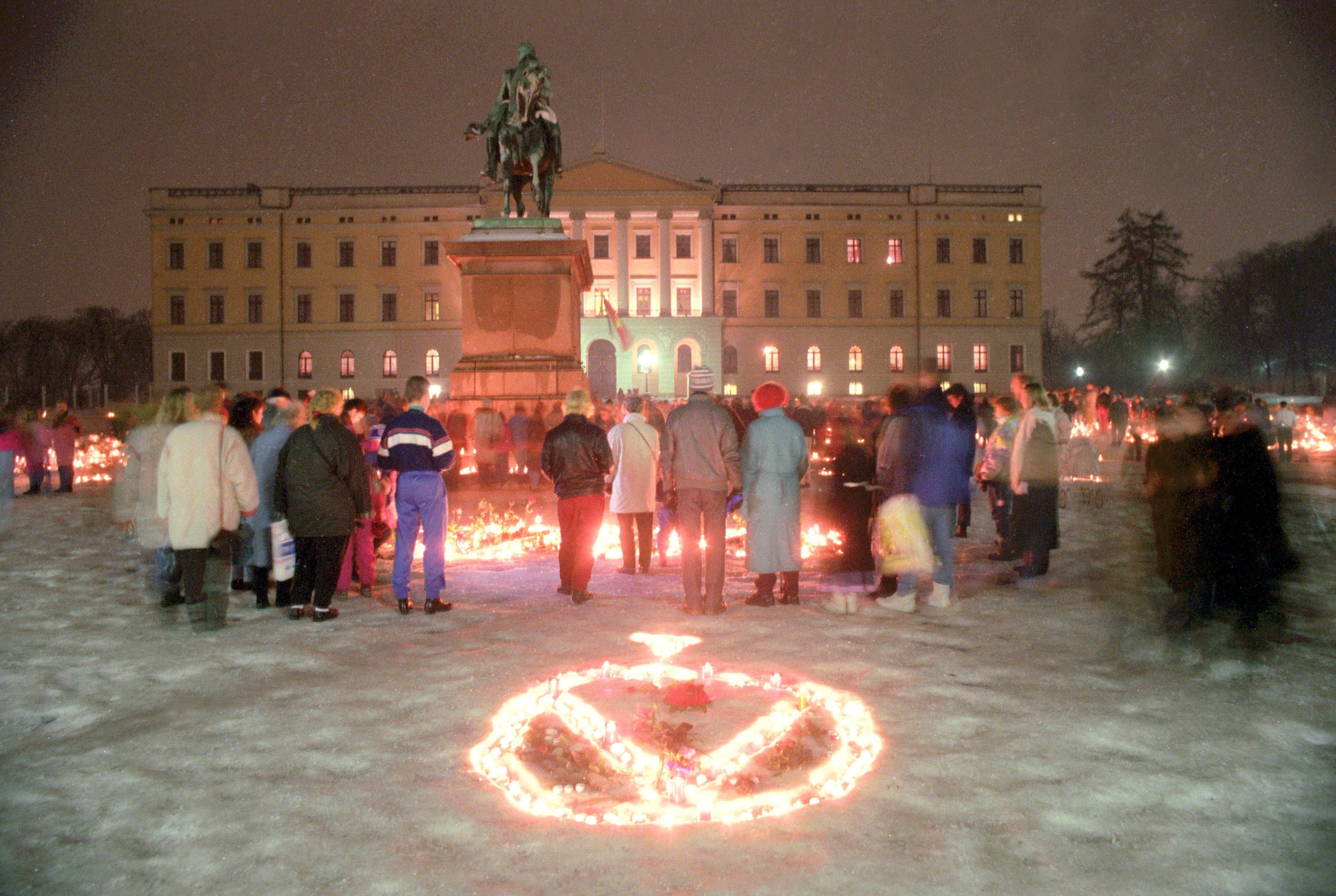 Slottsplassen full av blomster og lys som folk har plassert i snøen. Nærmest kameraet har noen laget Kong Olavs monogram med lys.