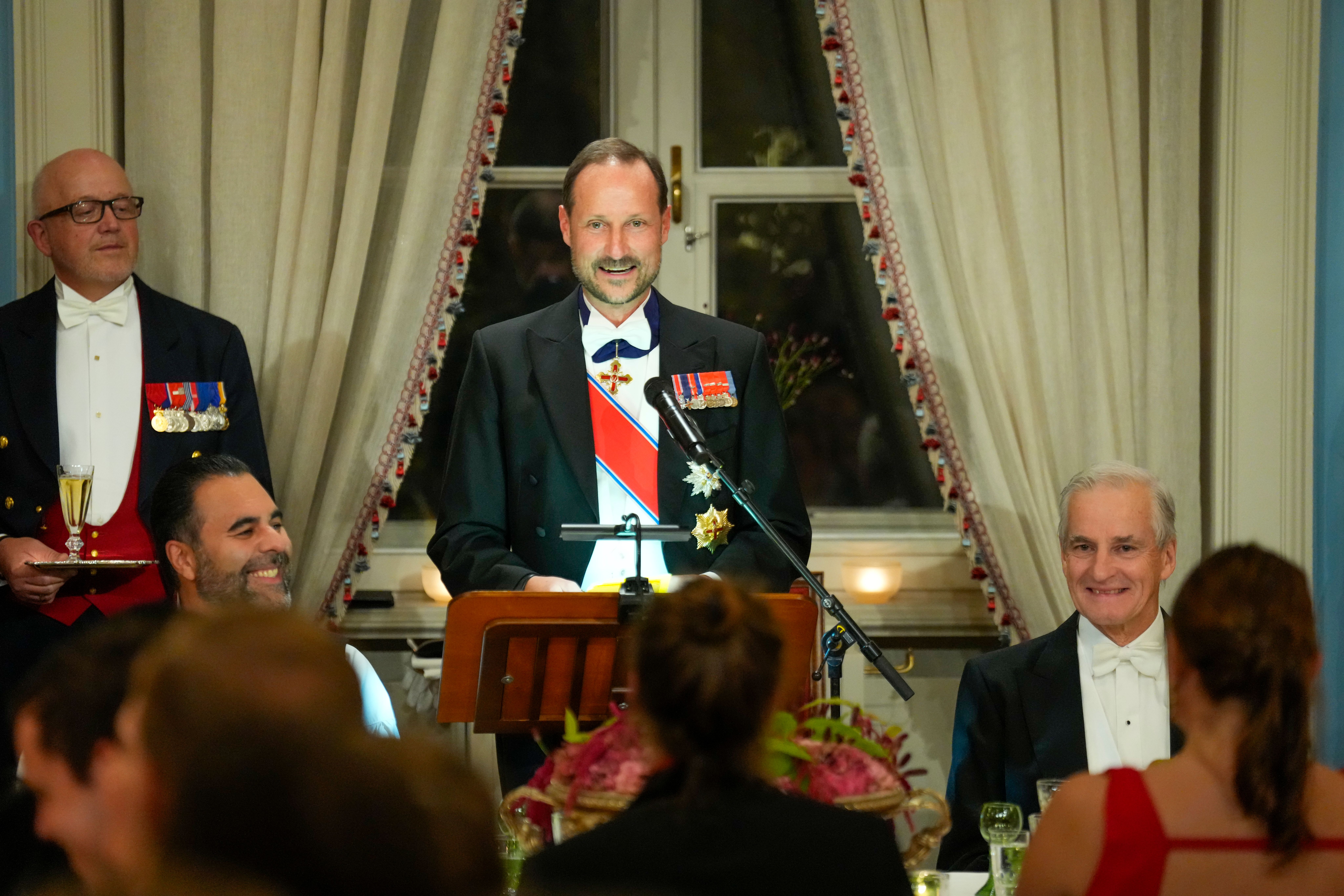 Kong Haakon står og holder tale. Stortingspresidenten og statsministeren sitter på hver sin side av ham, smiler og ler