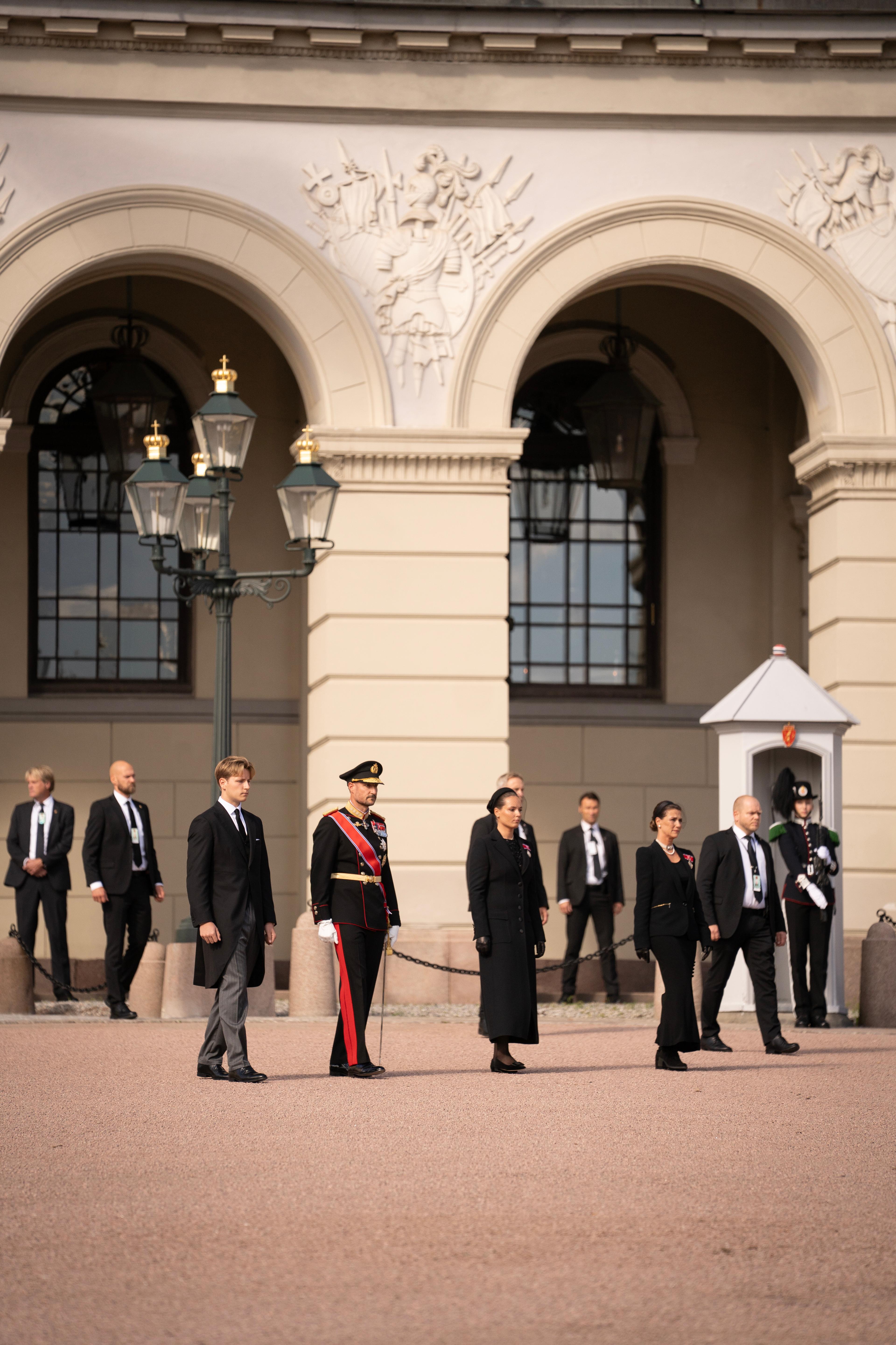 Prins Sverre Magnus, Kong Haakon, Kronprinsesse Ingrid Alexandra og Prinsesse Märtha Louise går over Slottsplassen. Slottet og gardist i skilderhus sees i bakgrunnen.