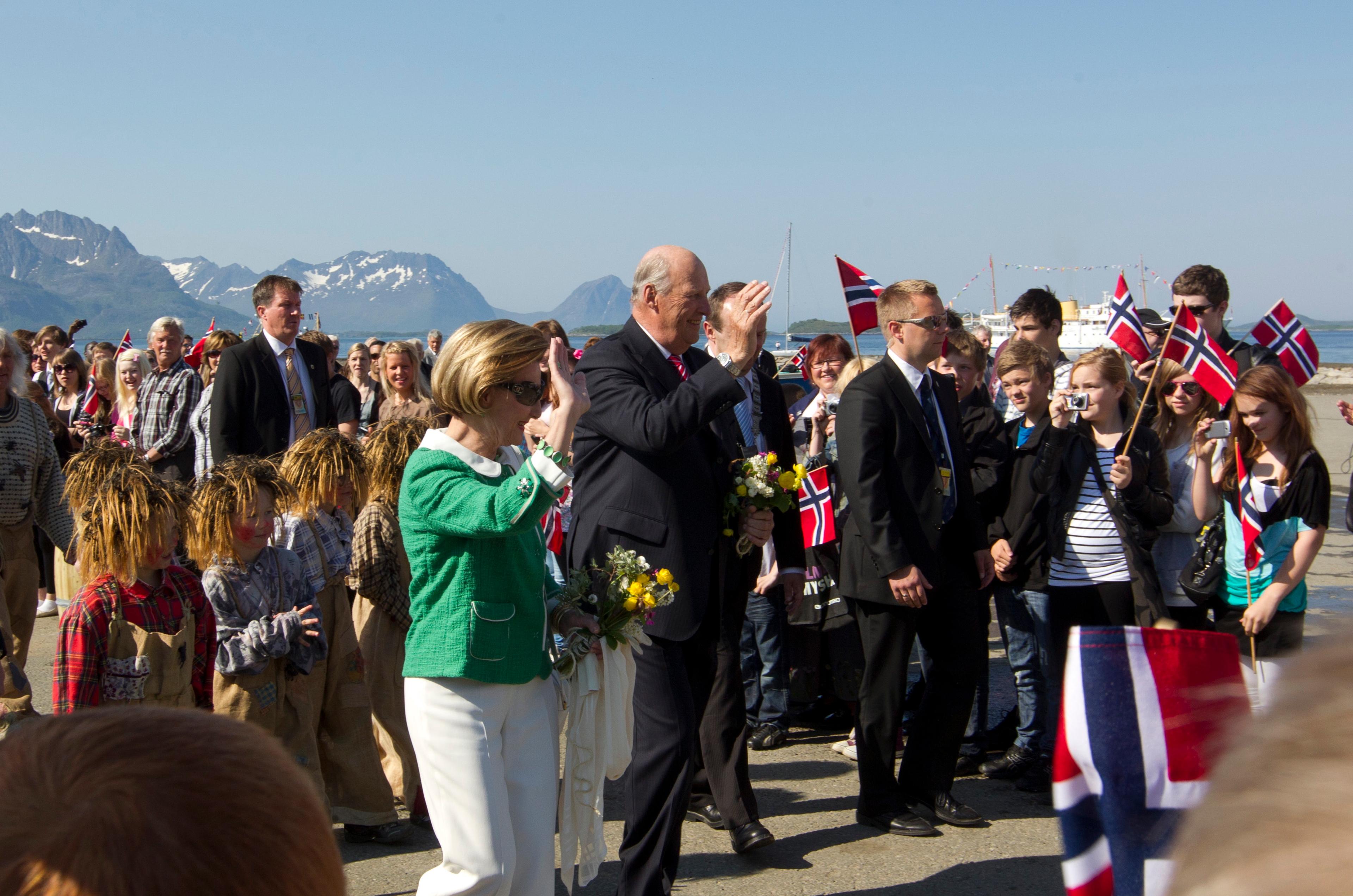 Kongeparet vinker til fremmøtte som flagger og vinker tilbake. Fjell vises i bakgrunnen