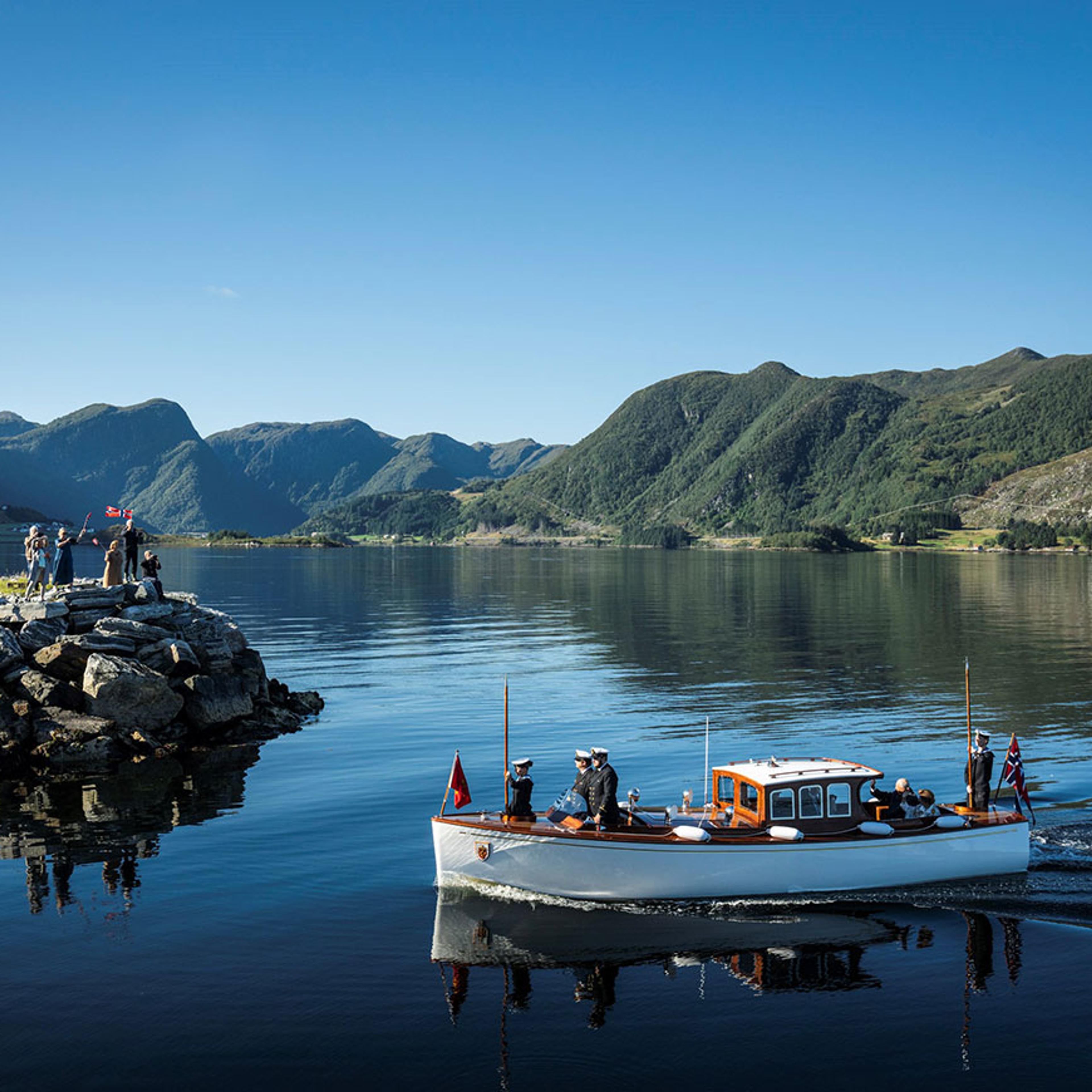 Sjaluppen fra Kongeskipet går gjennom speilbankt, blått vann inn mot Vanylven. Det er fjell i bakgrunnen og ute på neset de passerer står folk med flagg og vinker.