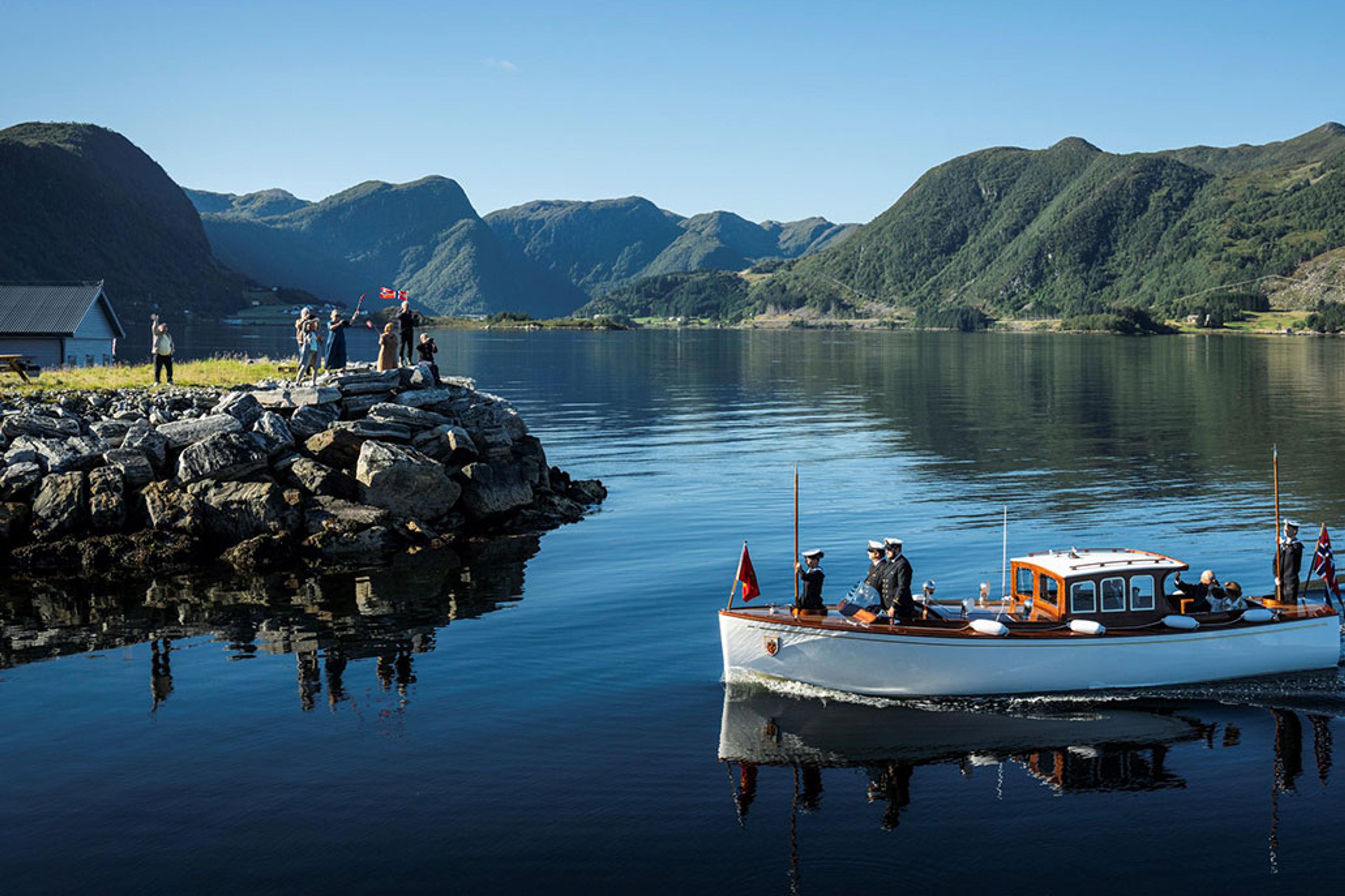 Sjaluppen fra Kongeskipet går gjennom speilbankt, blått vann inn mot Vanylven. Det er fjell i bakgrunnen og ute på neset de passerer står folk med flagg og vinker.