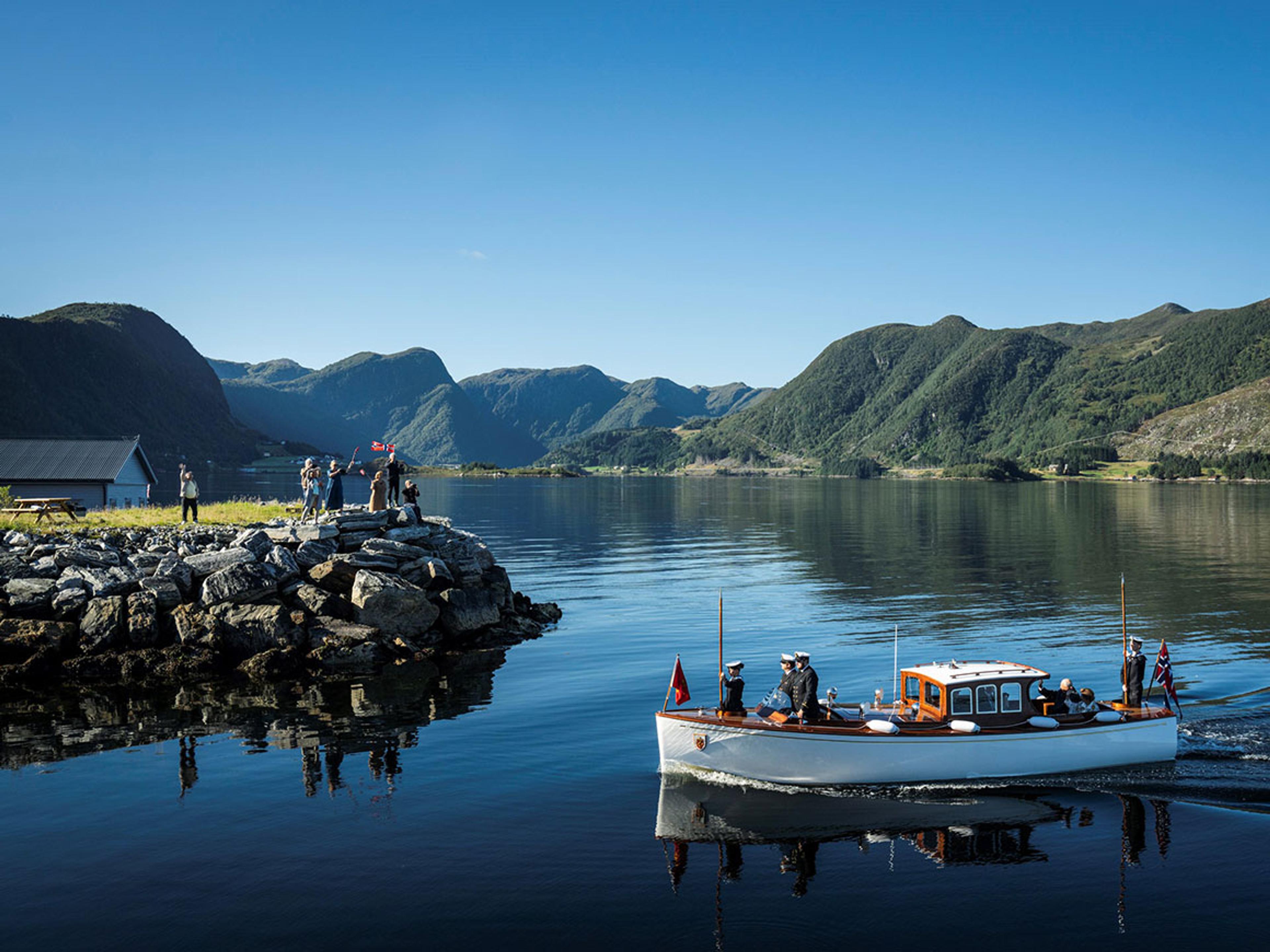Sjaluppen fra Kongeskipet går gjennom speilbankt, blått vann inn mot Vanylven. Det er fjell i bakgrunnen og ute på neset de passerer står folk med flagg og vinker.