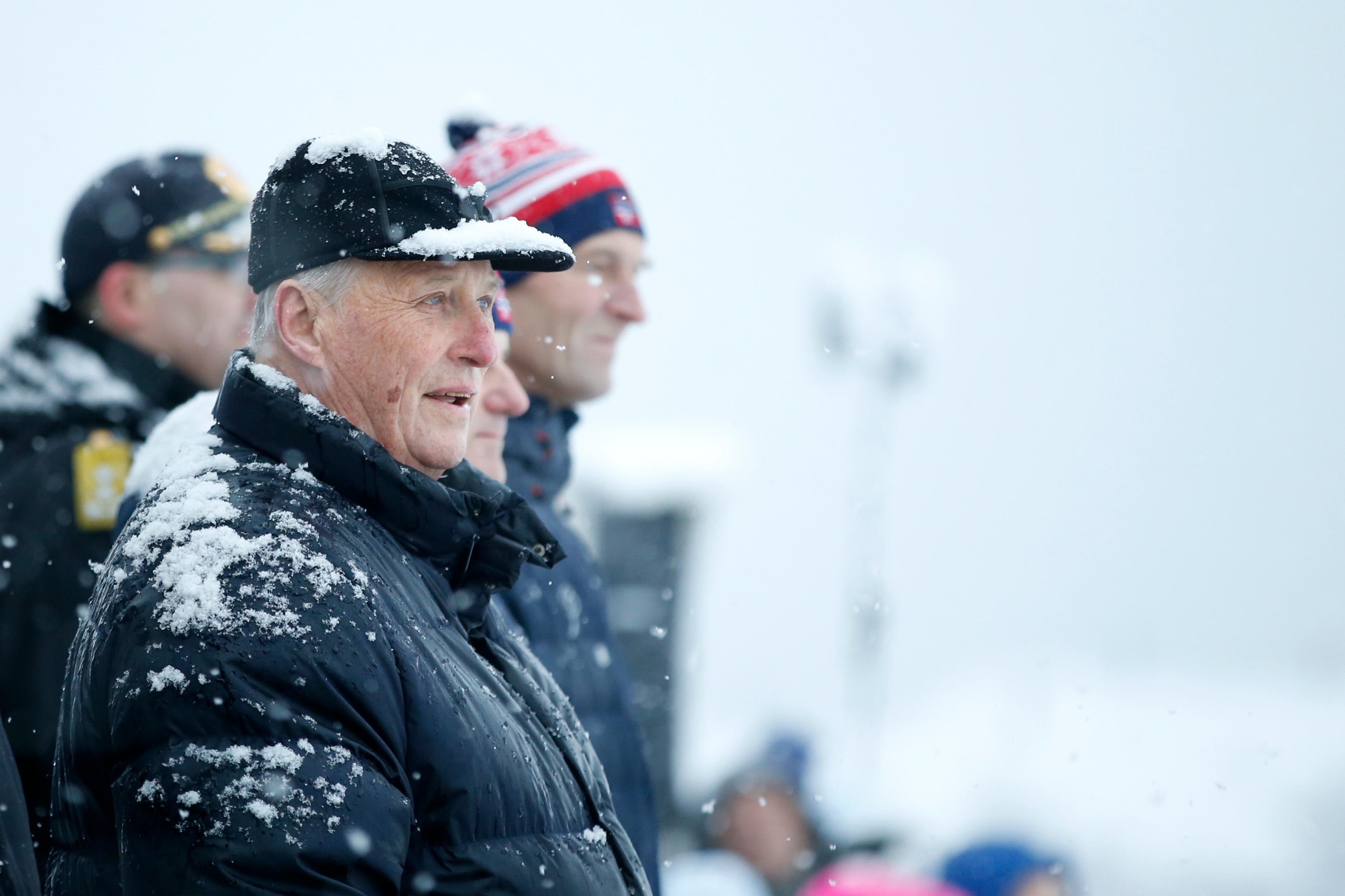 Kong Harald står på tribunen med masse snø, både på skyggelua og jakken sin.
