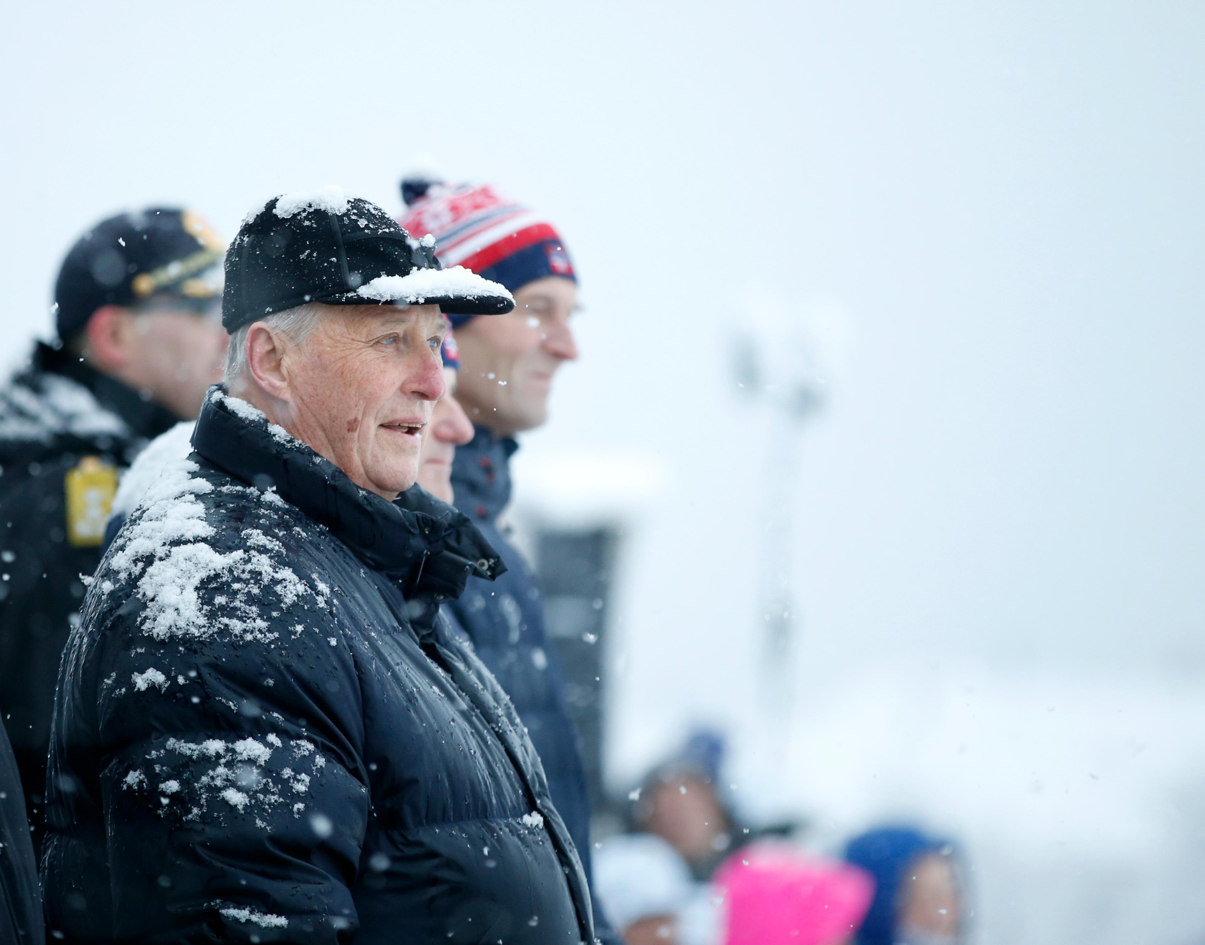 Kong Harald står på tribunen med masse snø, både på skyggelua og jakken sin.