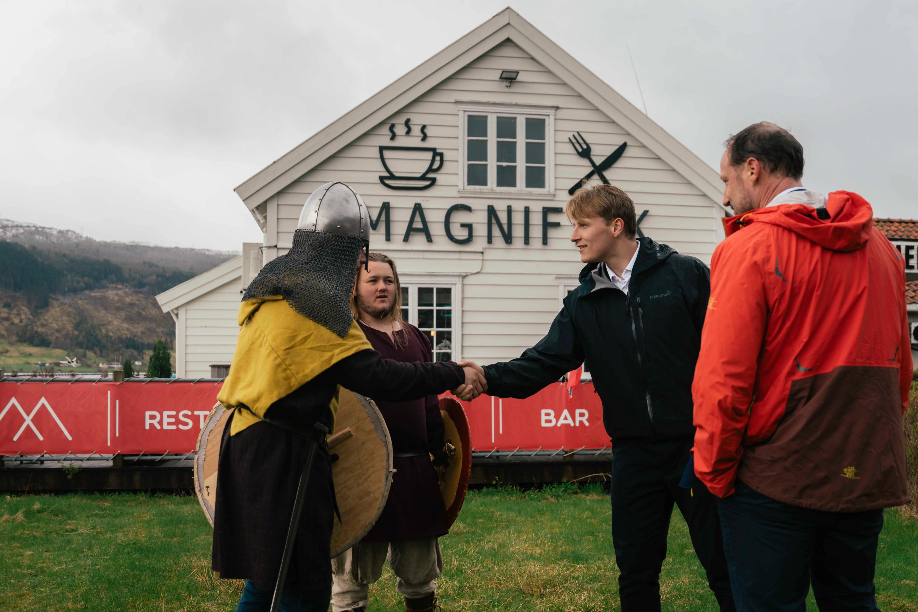 Prins Sverre Magnus strekker ut hånden og hilser på en person kledd i vikingklær med hjelm og skjold. På bildet er også Kronprins Haakon og en til person kledd i vikingantrekk. I bakgunnen ligger det en café