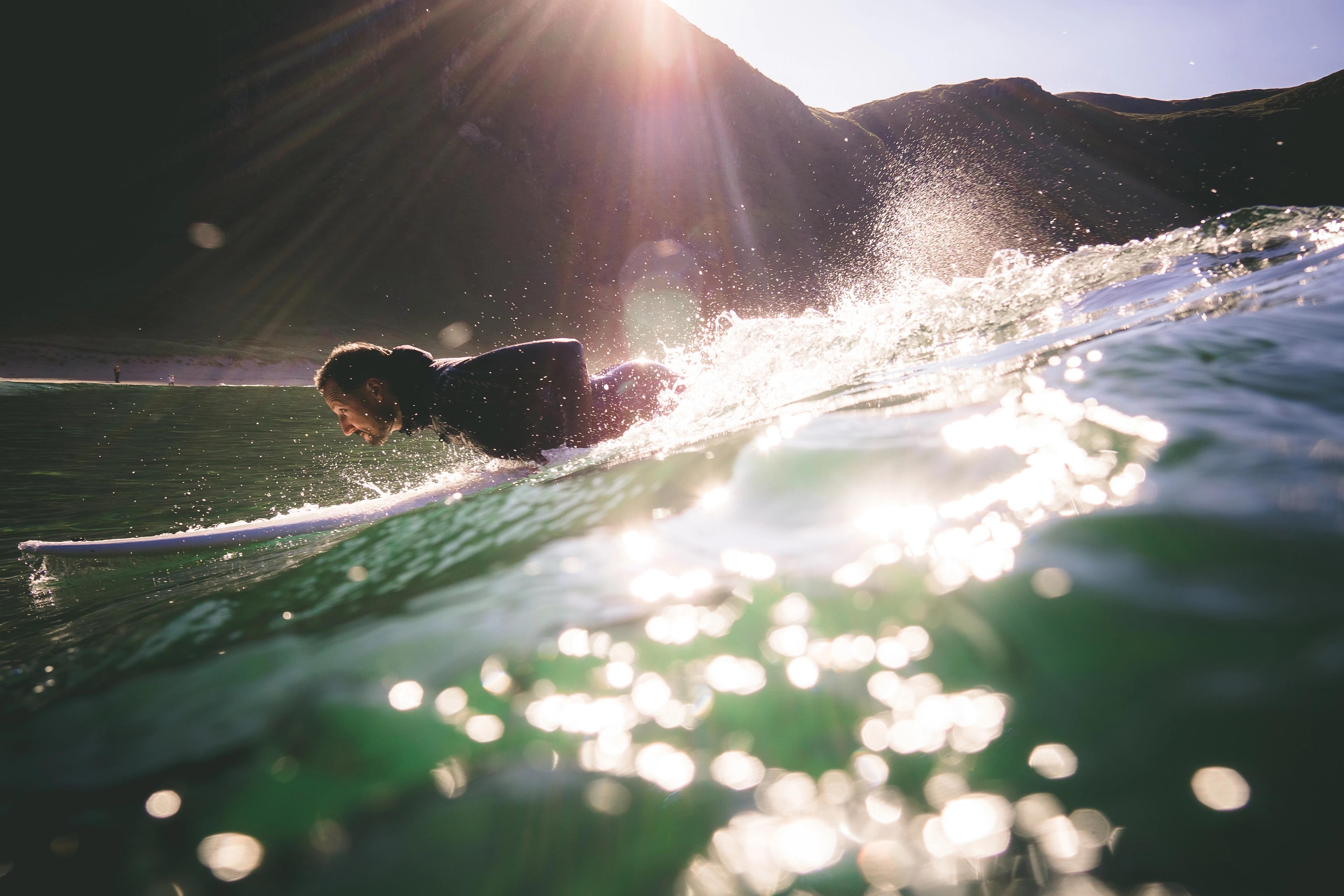 Kronprins Haakon liggende forover på surfebrettet med solskimmer på bølgene