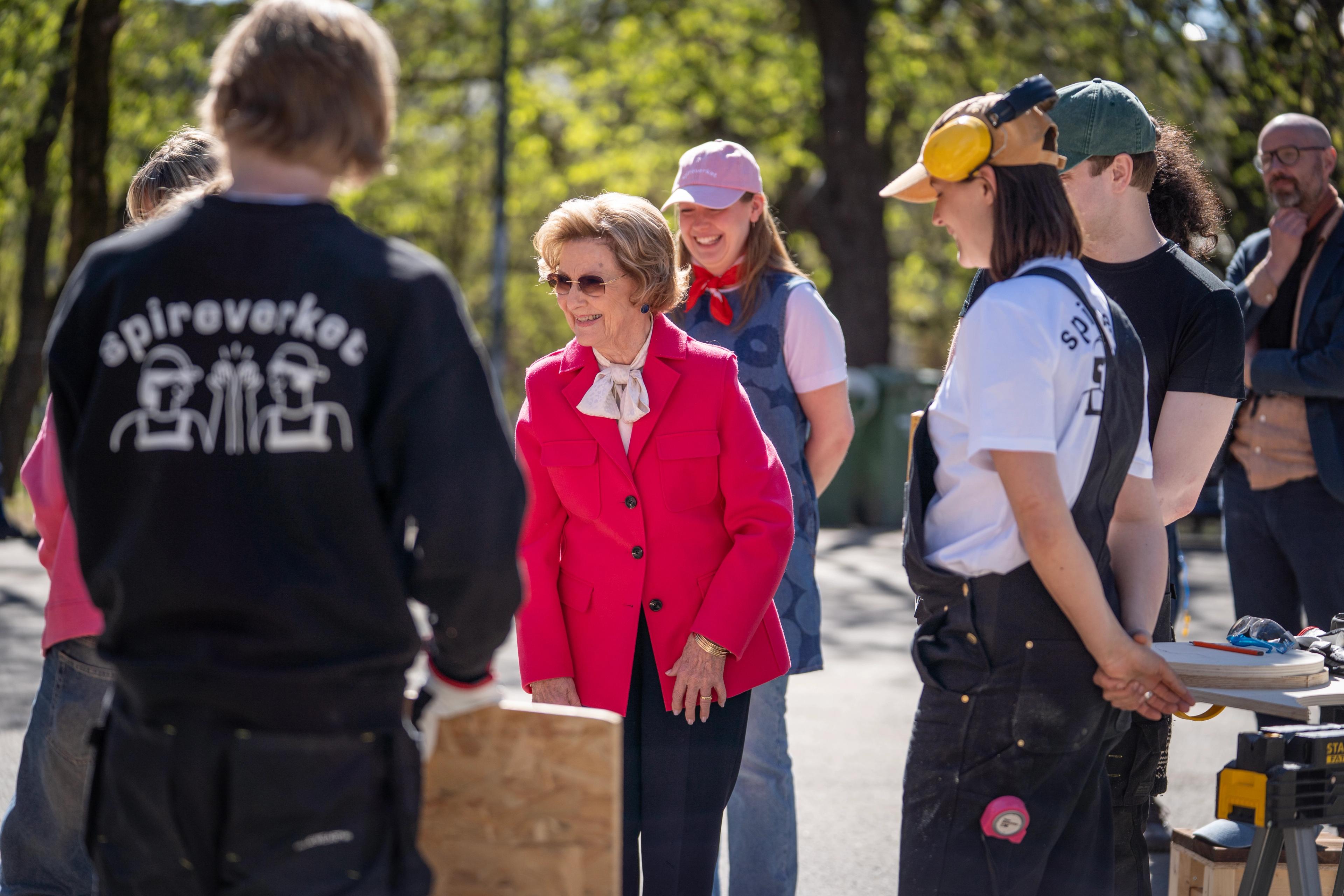 Dronning Sonja står i en ring med unge voksne i arbeidsantrekk for møbelsnekring.
