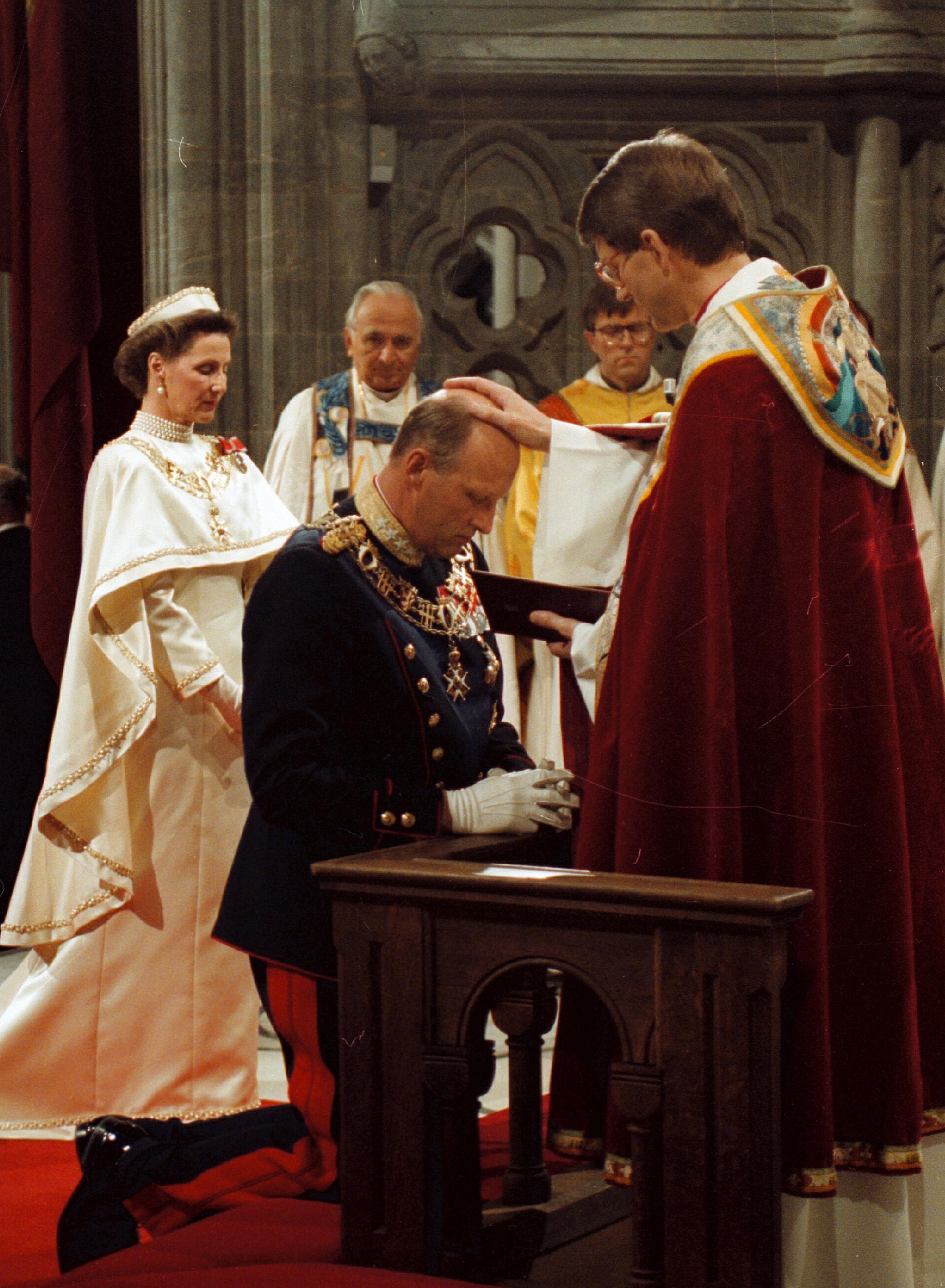 Kong Harald kneler foran biskopen, som holder hånden på Kongens hode. Biskopen har rød kappe, Kongen er i gallauniform. Dronning Sonja står i bakgrunnen i sin hvite kroningskjole.
