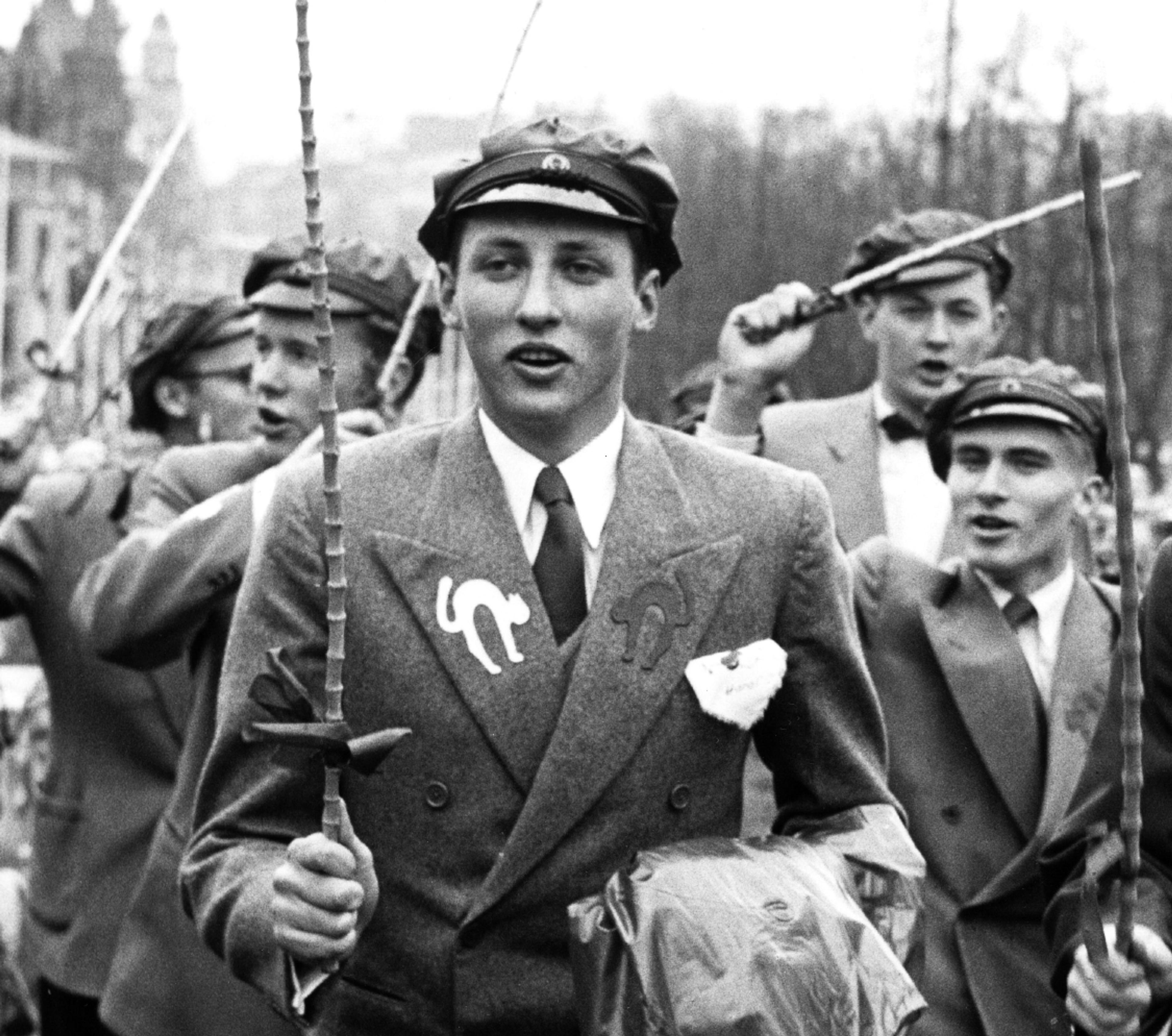 Sort-hvittbilde av Prins Harald i russetoget. Han har russelue og russestokk, skjorte og slips. Jakken har en sort og en hvit katt applikert på slagene.