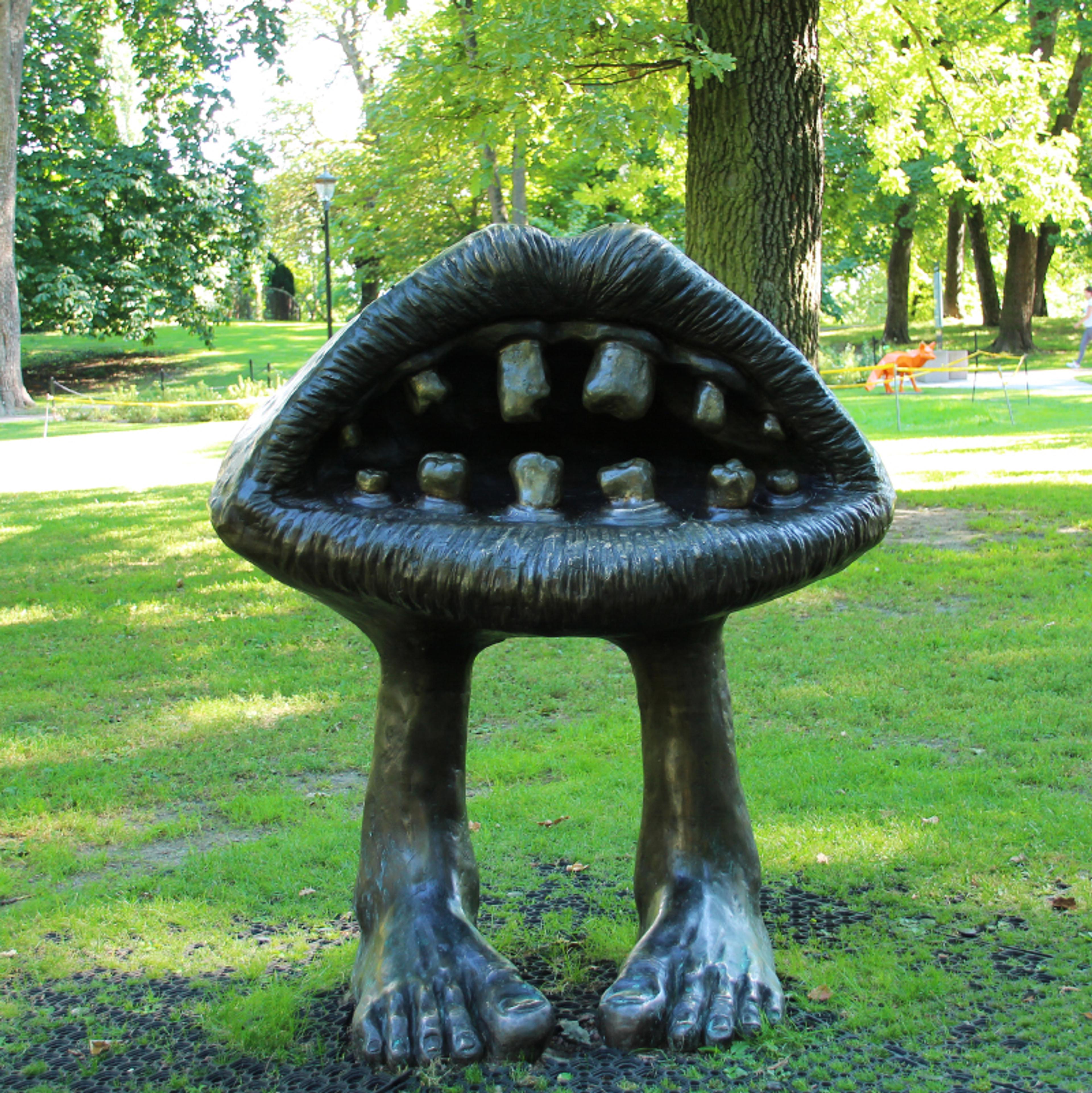 En stor munn, støpt i mørkt metall, står åpen med lepper og tenner står på egne ben i gresset.
