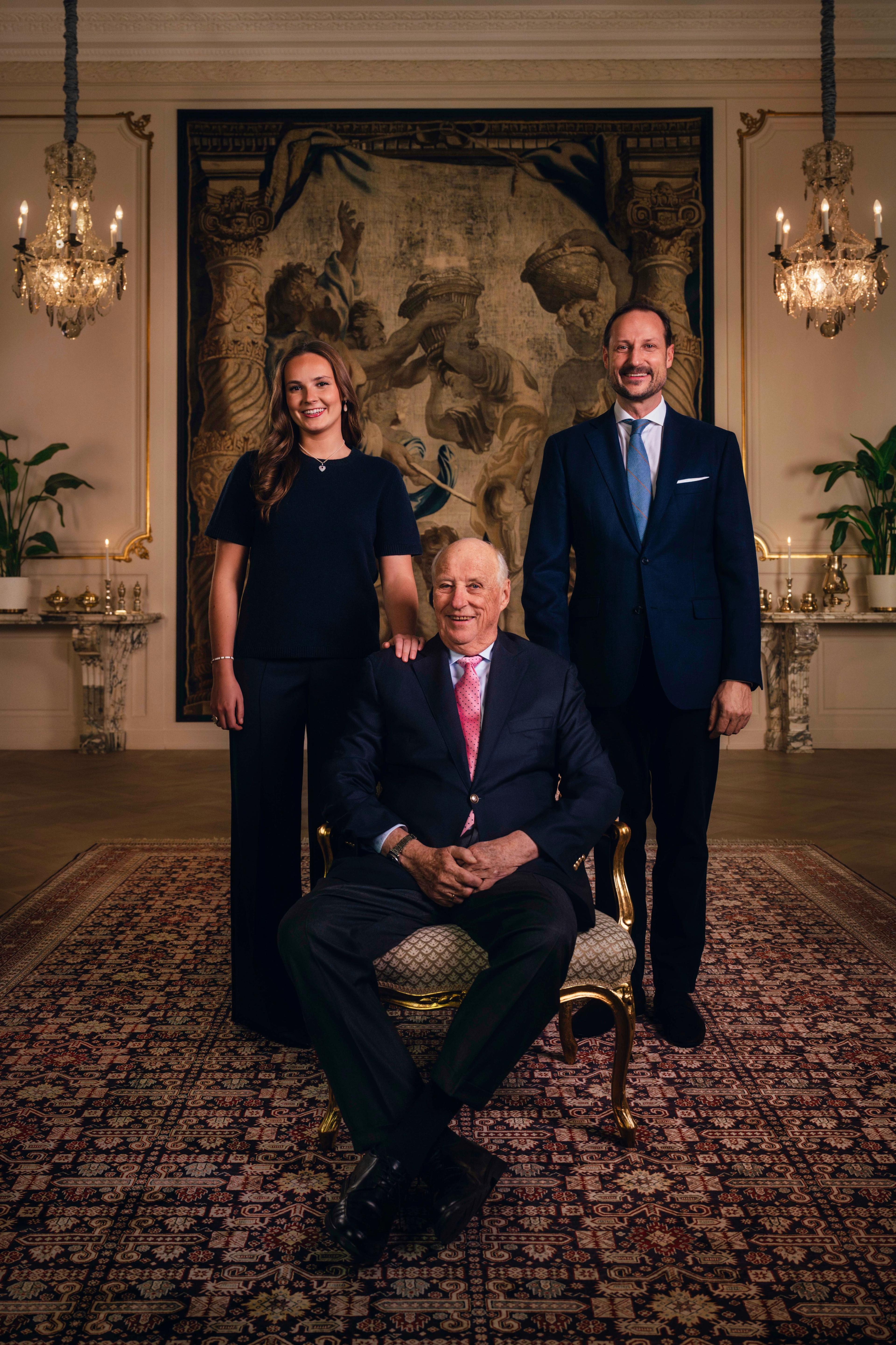 Kong Harald sittende i en stol, flankert av Kronprins Haakon og Prinsesse Ingrid Alexandra.