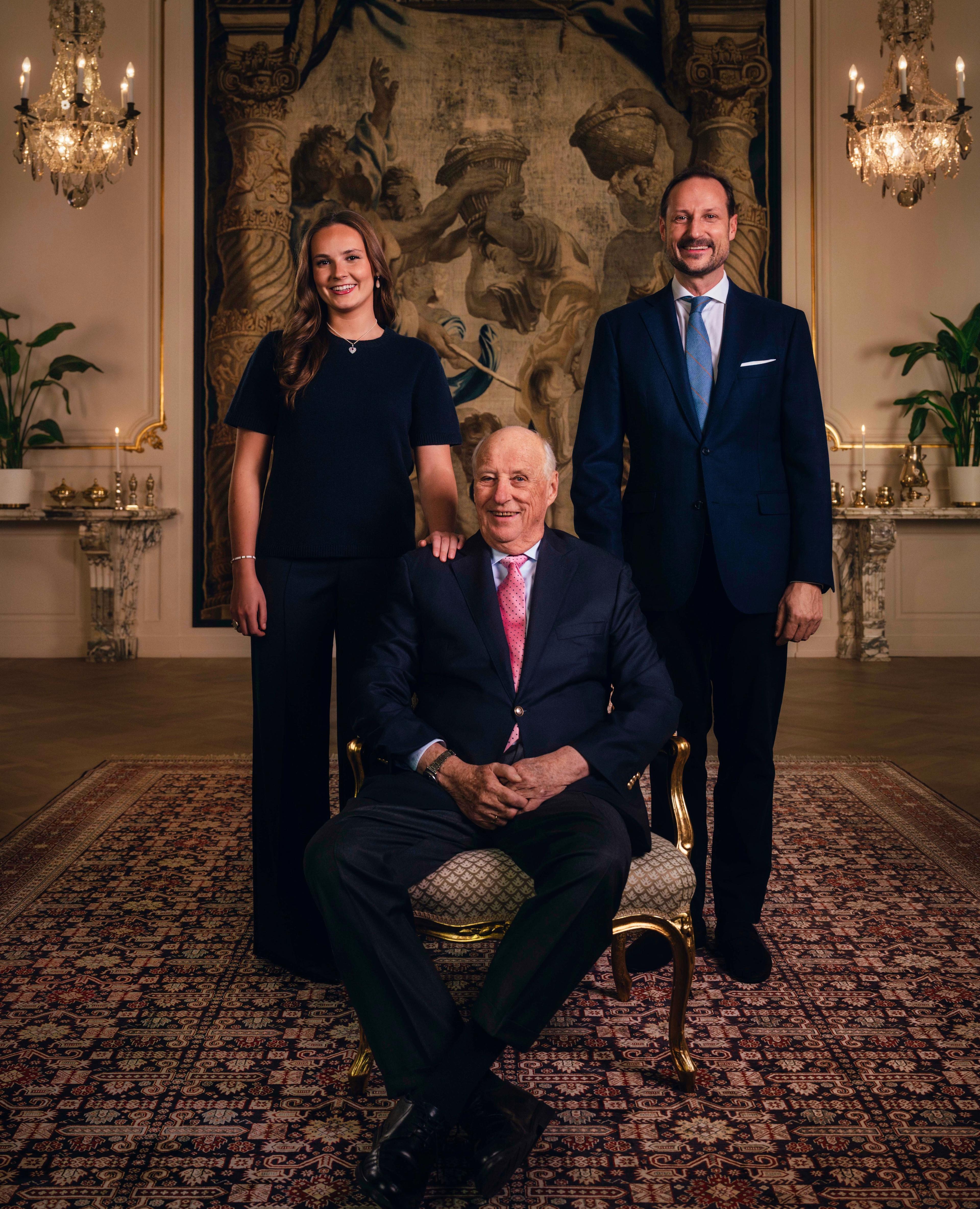 Kong Harald sittende i en stol, flankert av Kronprins Haakon og Prinsesse Ingrid Alexandra.