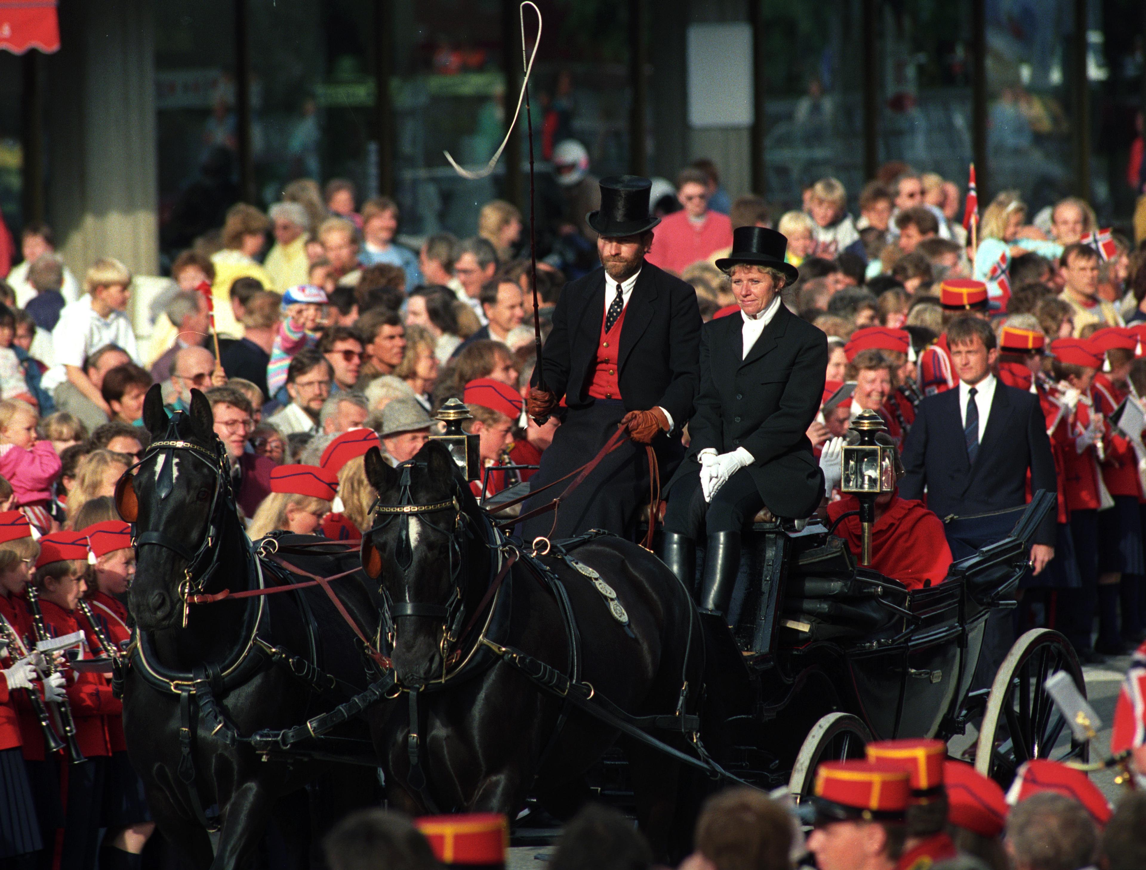 To svarte hester med hvitt i pannene trekker en åpen vogn. På bukken sitter kuskene med sorte flosshatter. Mange mennesker omgir vognen.