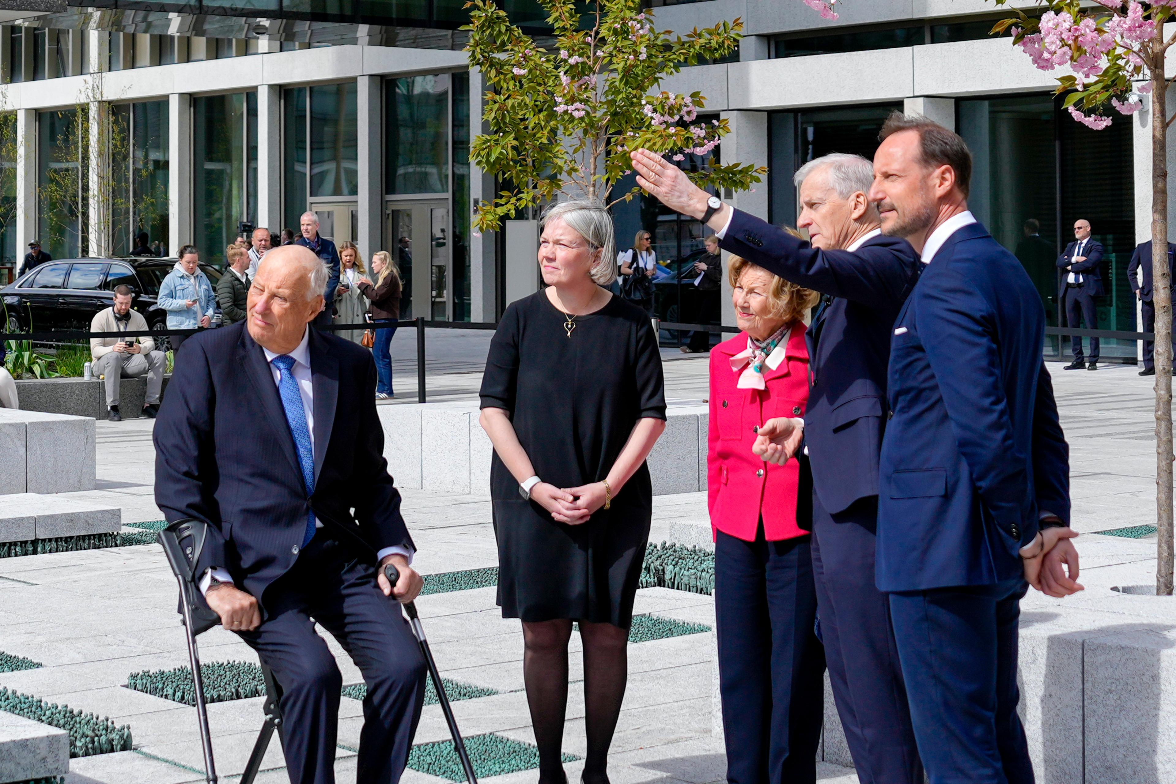 Kongen, Dronningen og Kronprinsen står sammen med statsministeren på plassen utenfor Høyblokka. Statsministeren peker mot noe utenfor bildet.