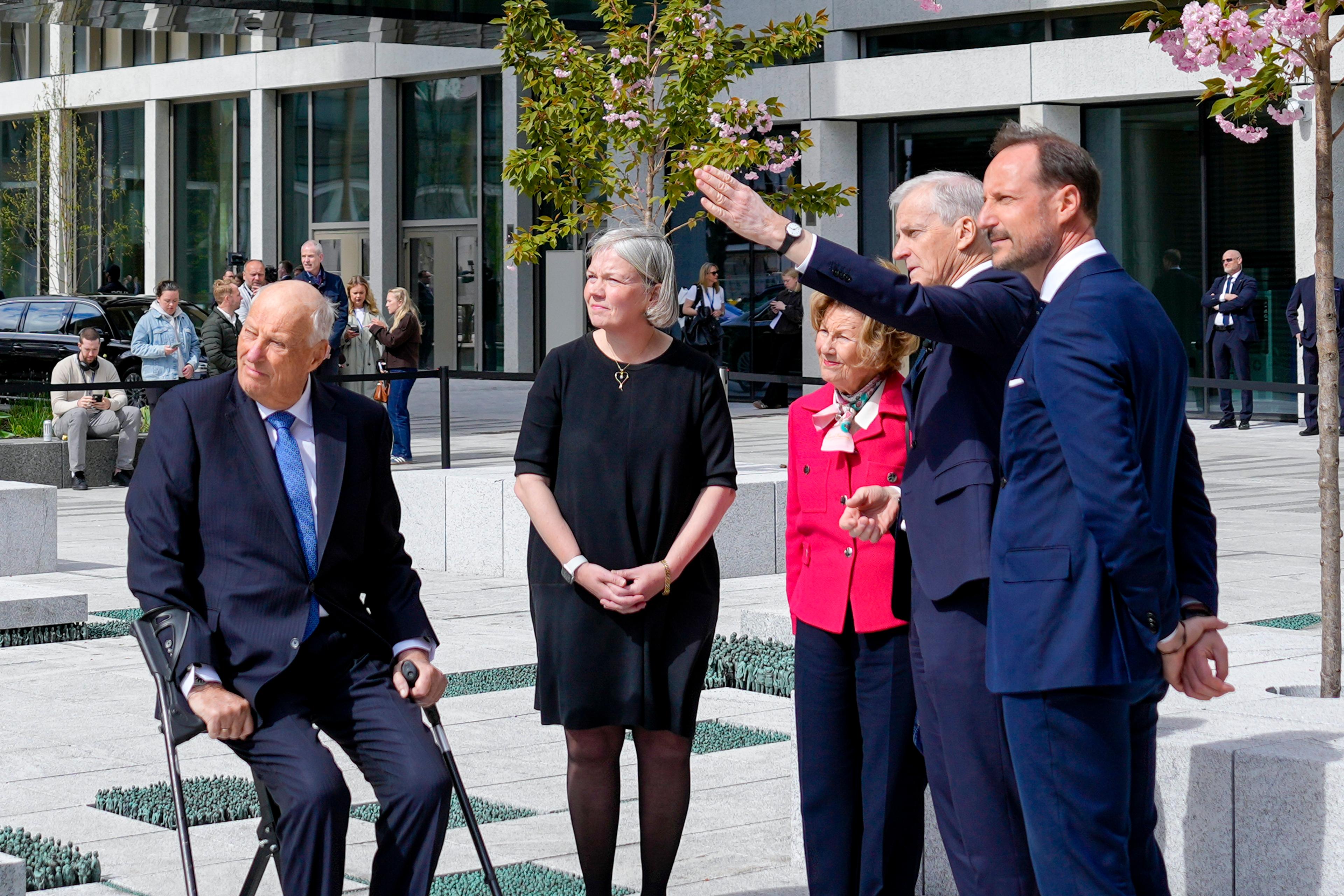 Kongen, Dronningen og Kronprinsen står sammen med statsministeren på plassen utenfor Høyblokka. Statsministeren peker mot noe utenfor bildet.