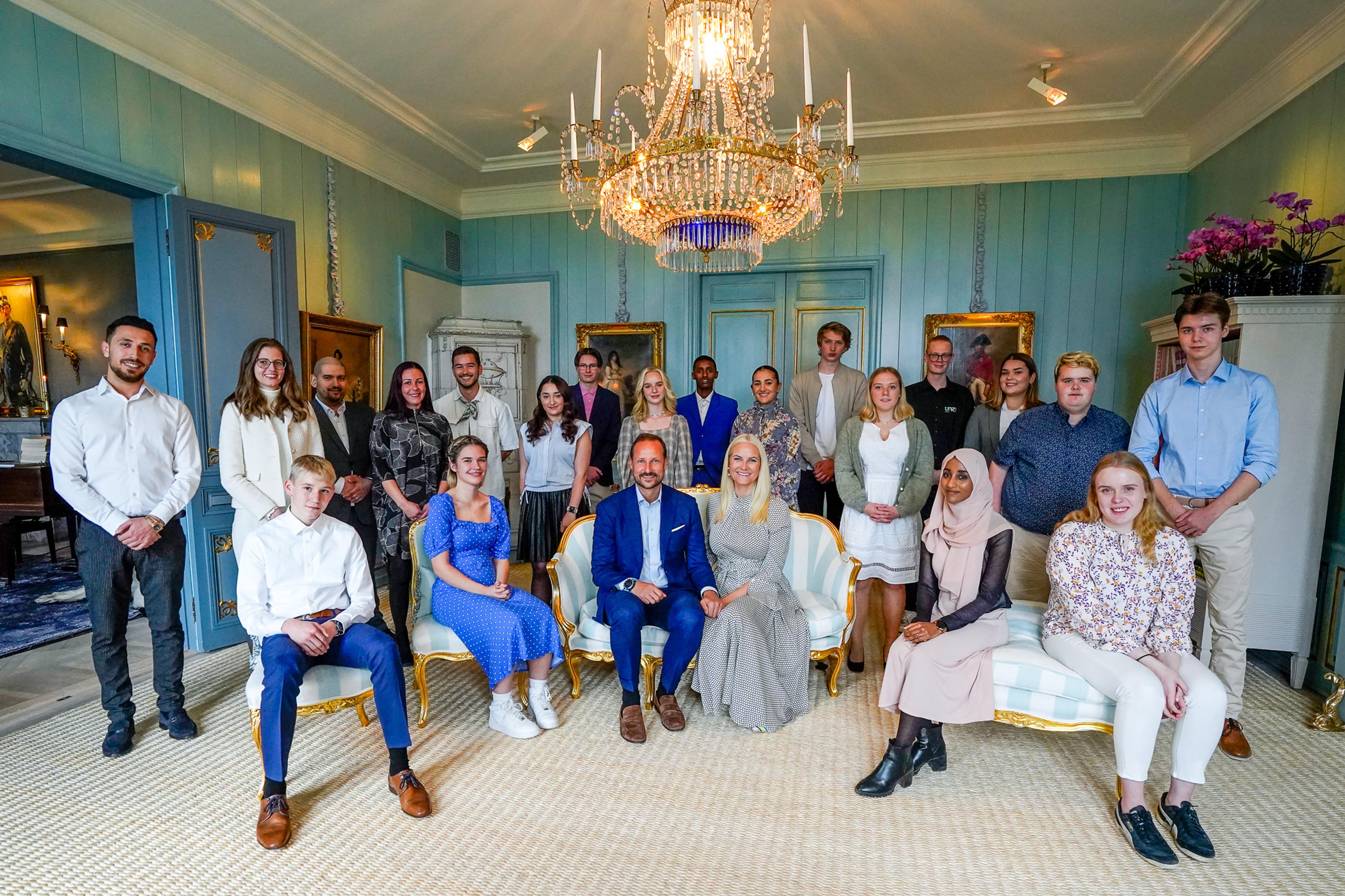 Kronprinsparet sitter i en liten sofa, omgitt av 20 unge mennesker som enten står eller sitter.