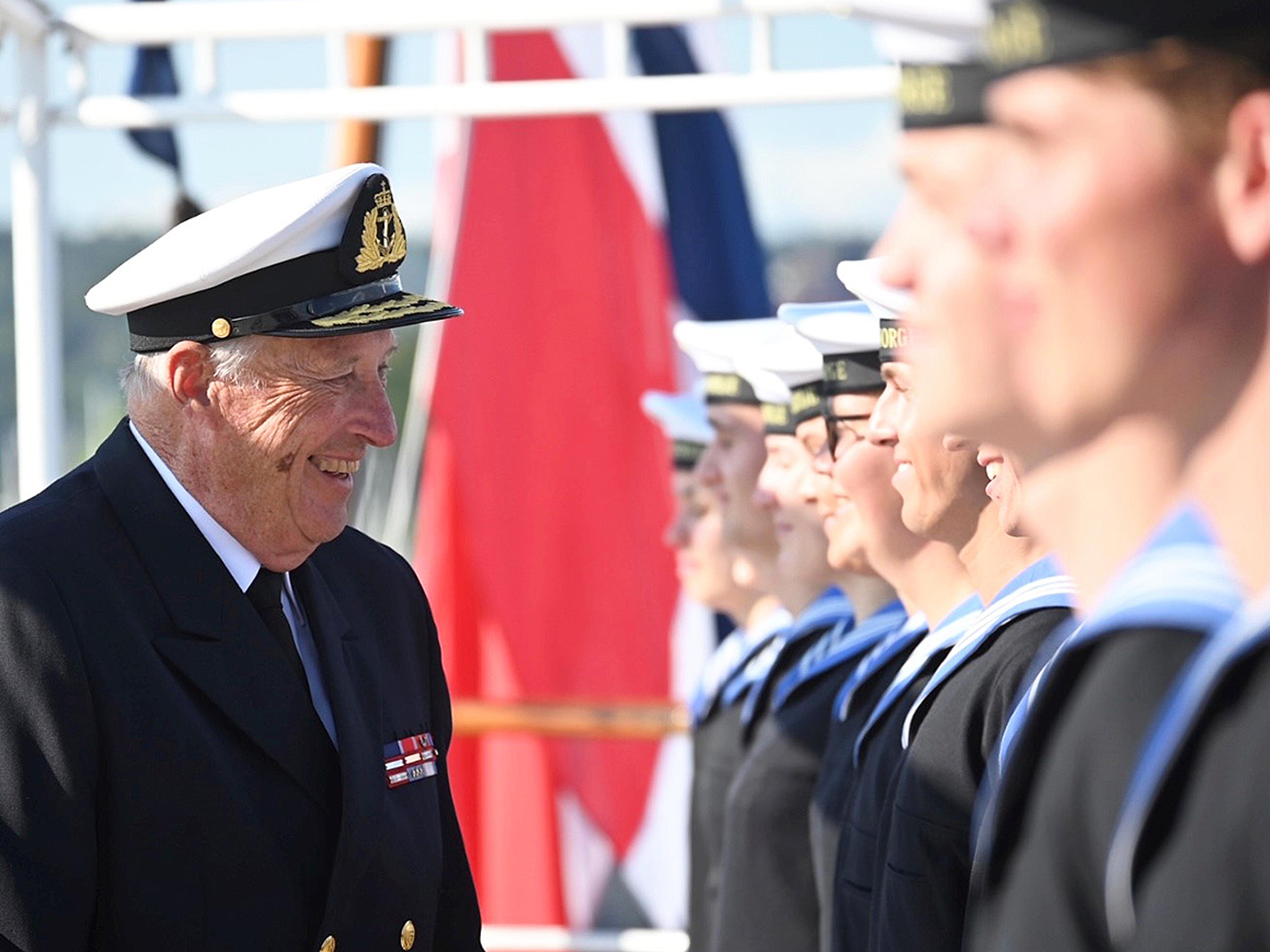 En smilende Kong Harald i marinens uniform snakker med gaster som står oppstilt for inspeksjon.