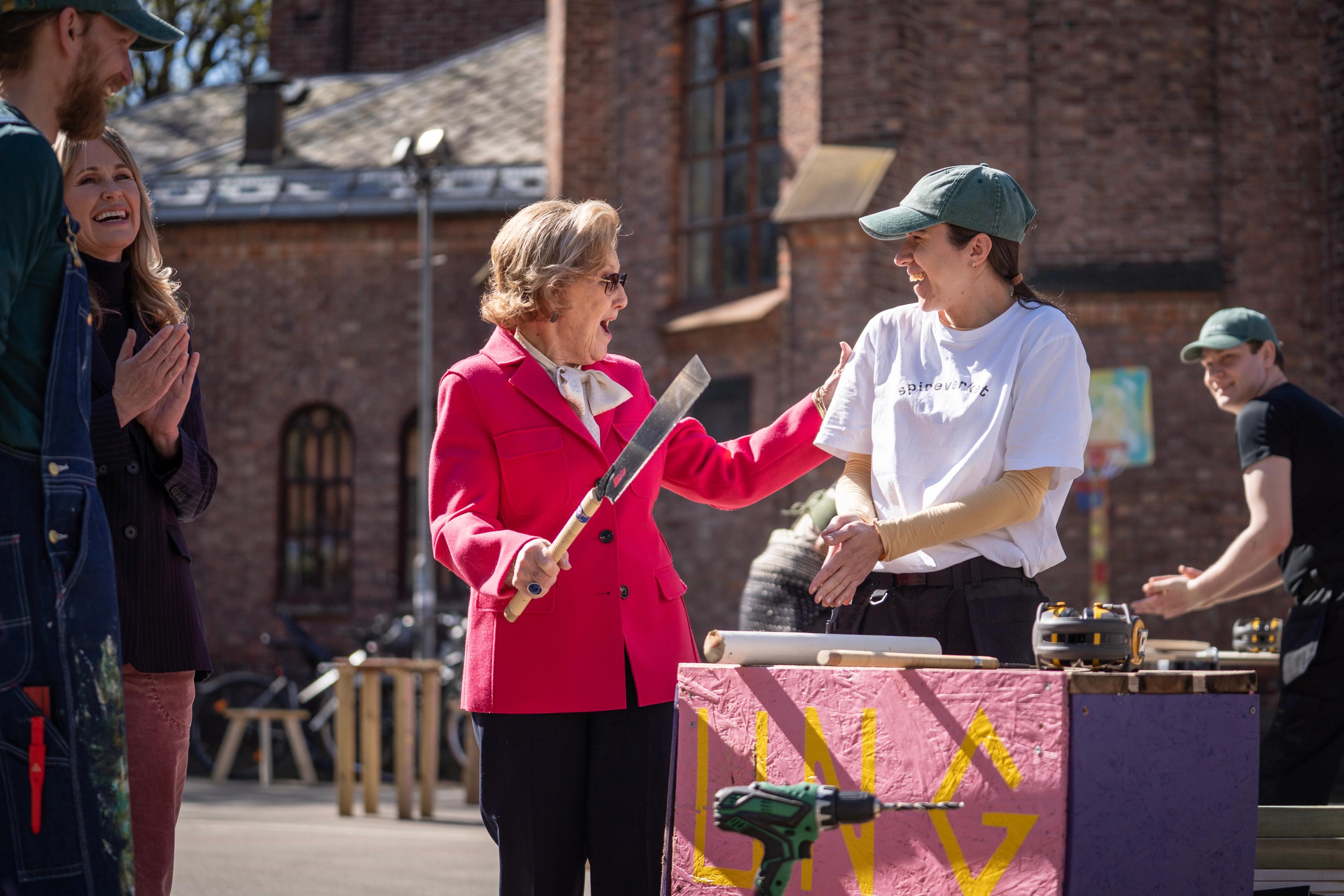 Dronning Sonja står med en sag i hånden og smiler bredt til en ansatt i Spireverket.