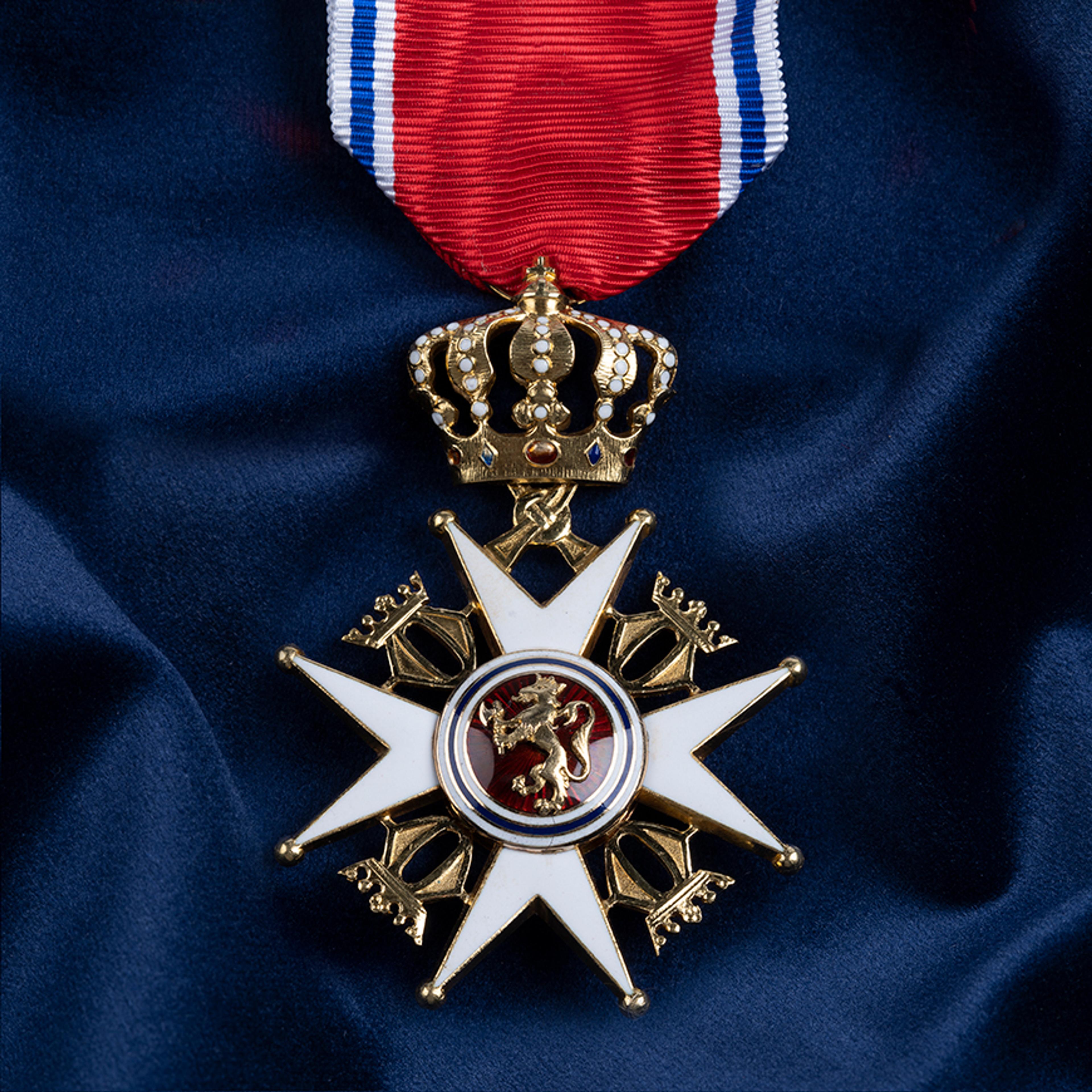 Nærbilde av ordensmerket festet i rødt bånd med blå og hvite striper på en bakgrunn av blått klede.
