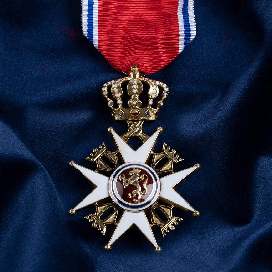 Nærbilde av ordensmerket festet i rødt bånd med blå og hvite striper på en bakgrunn av blått klede.