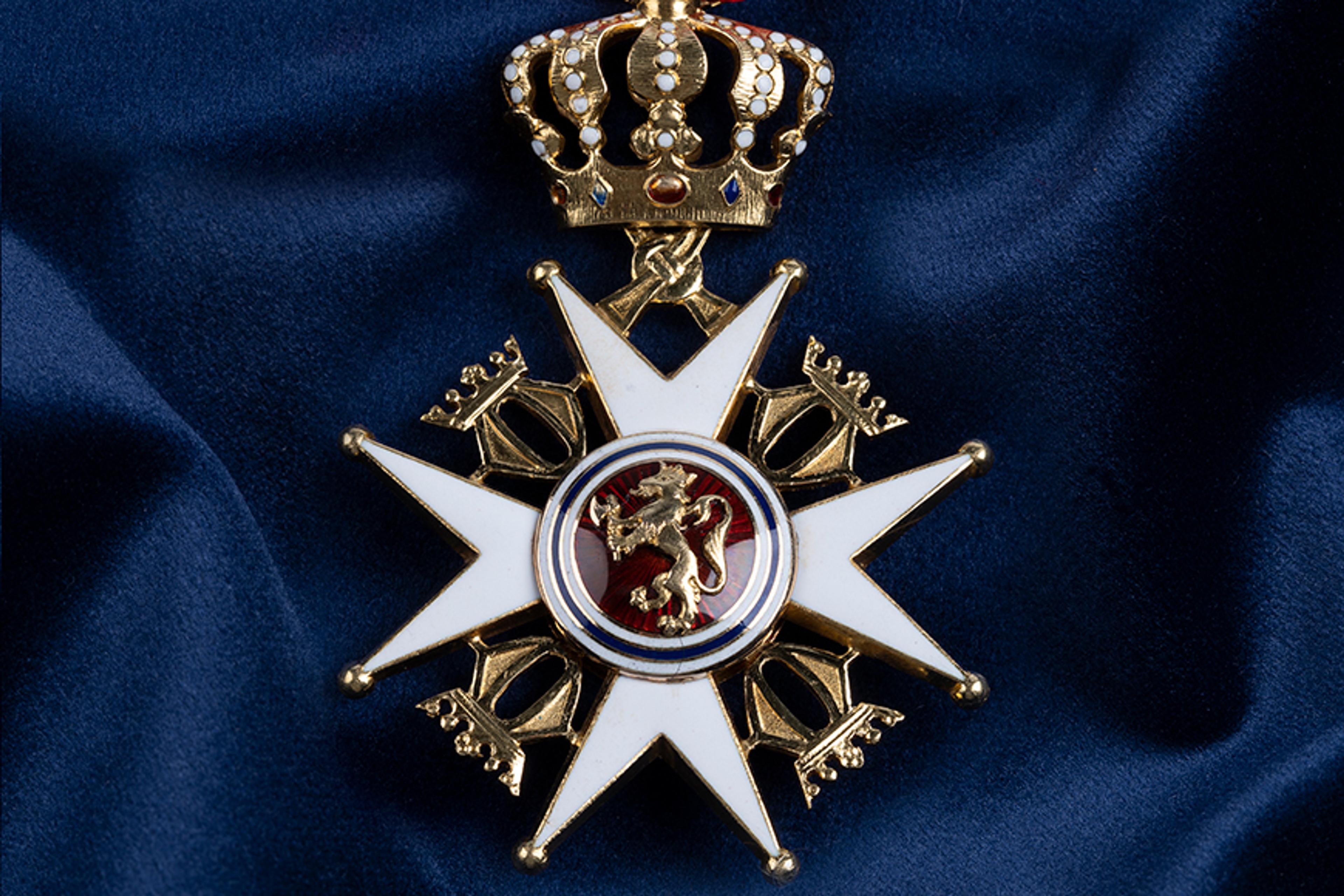 Nærbilde av ordensmerket festet i rødt bånd med blå og hvite striper på en bakgrunn av blått klede.