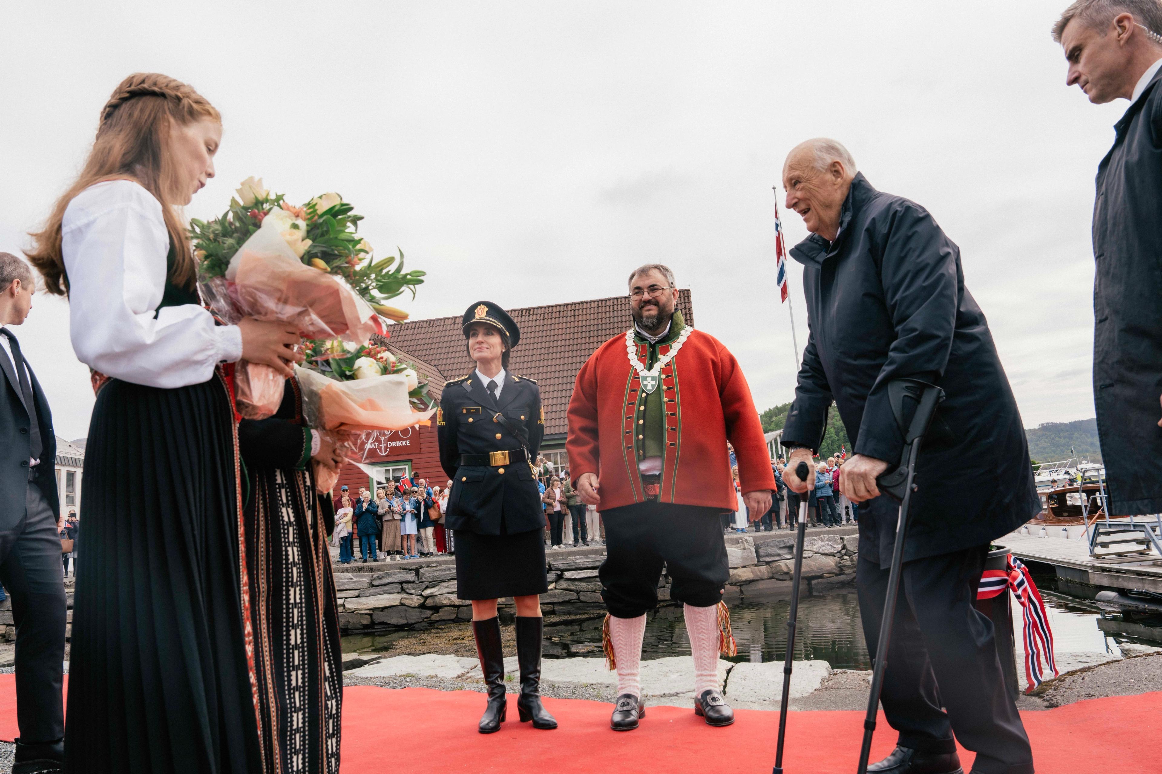 Kong Harald står på kaia, omringa av ordførar, politimeister og ei jente med blomar.