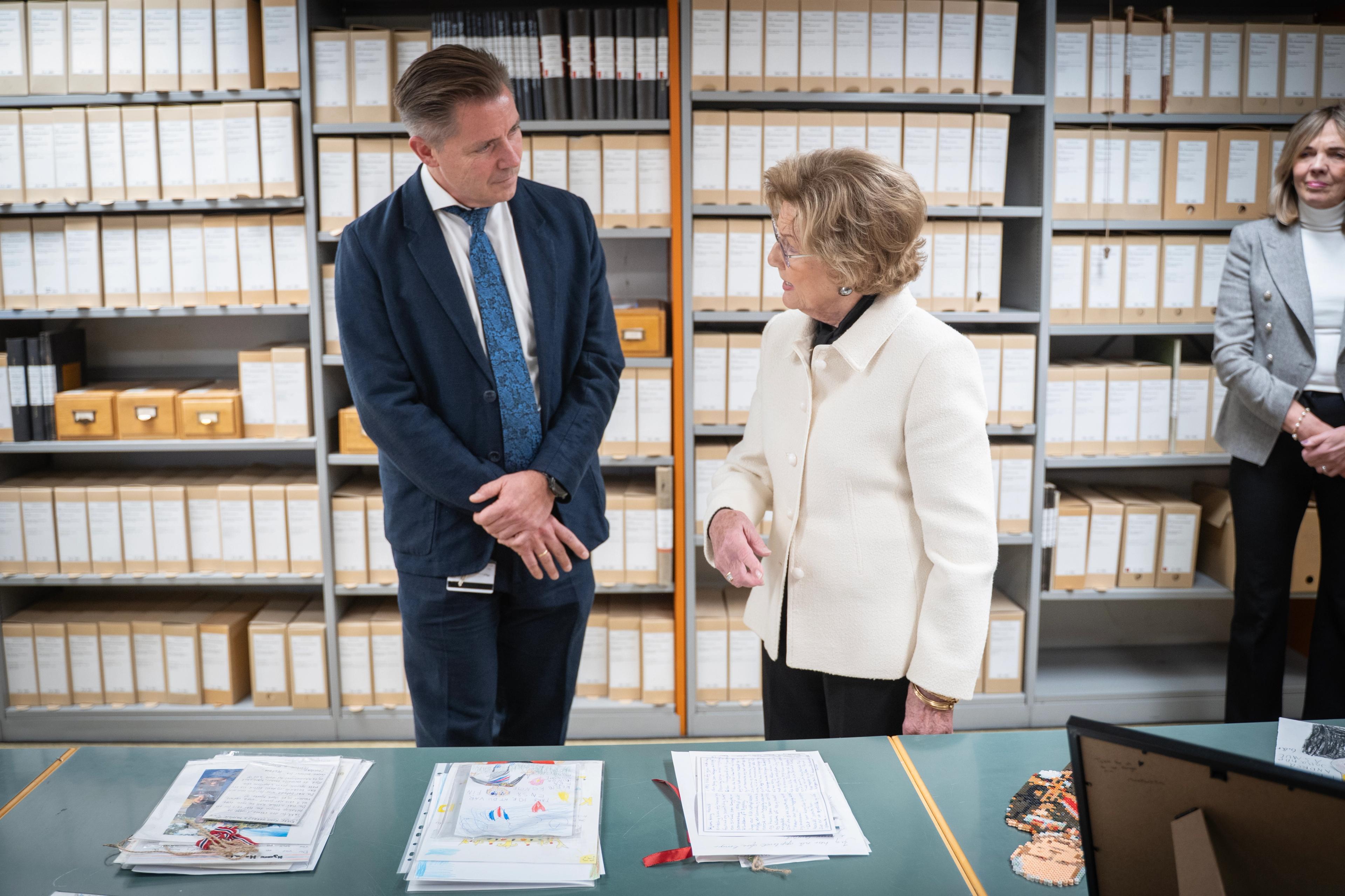 Dronning Sonja peker på bordet med brev og hilsener, mens hun ser på Stian Nordli. Bak er hyller med ulike kartonger.