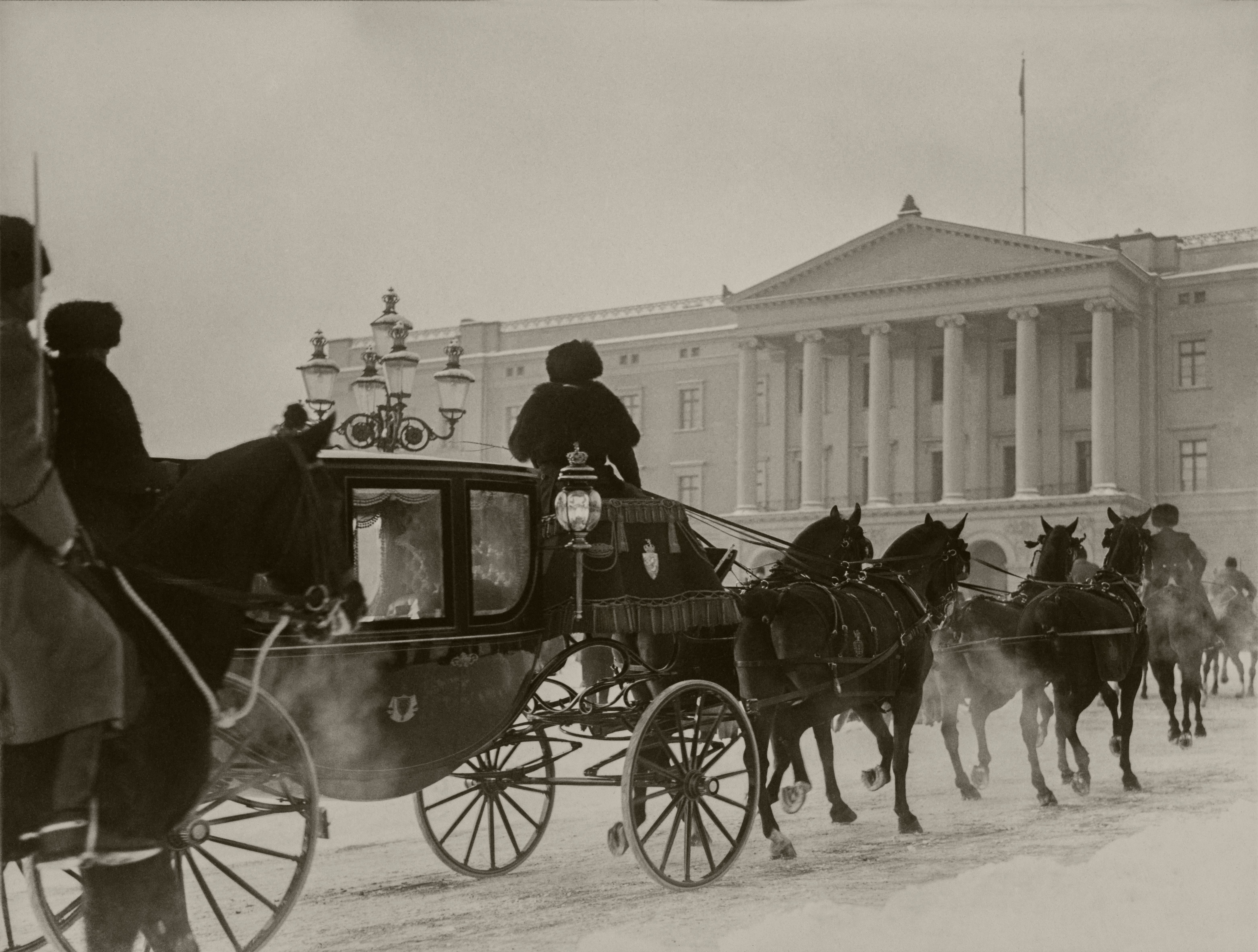 Sort-hvittbilde av vognen trukket av fire mørke hester på vei opp den snølagte bakken inn på Slottsplassen. Slottets fasade i foran dem.