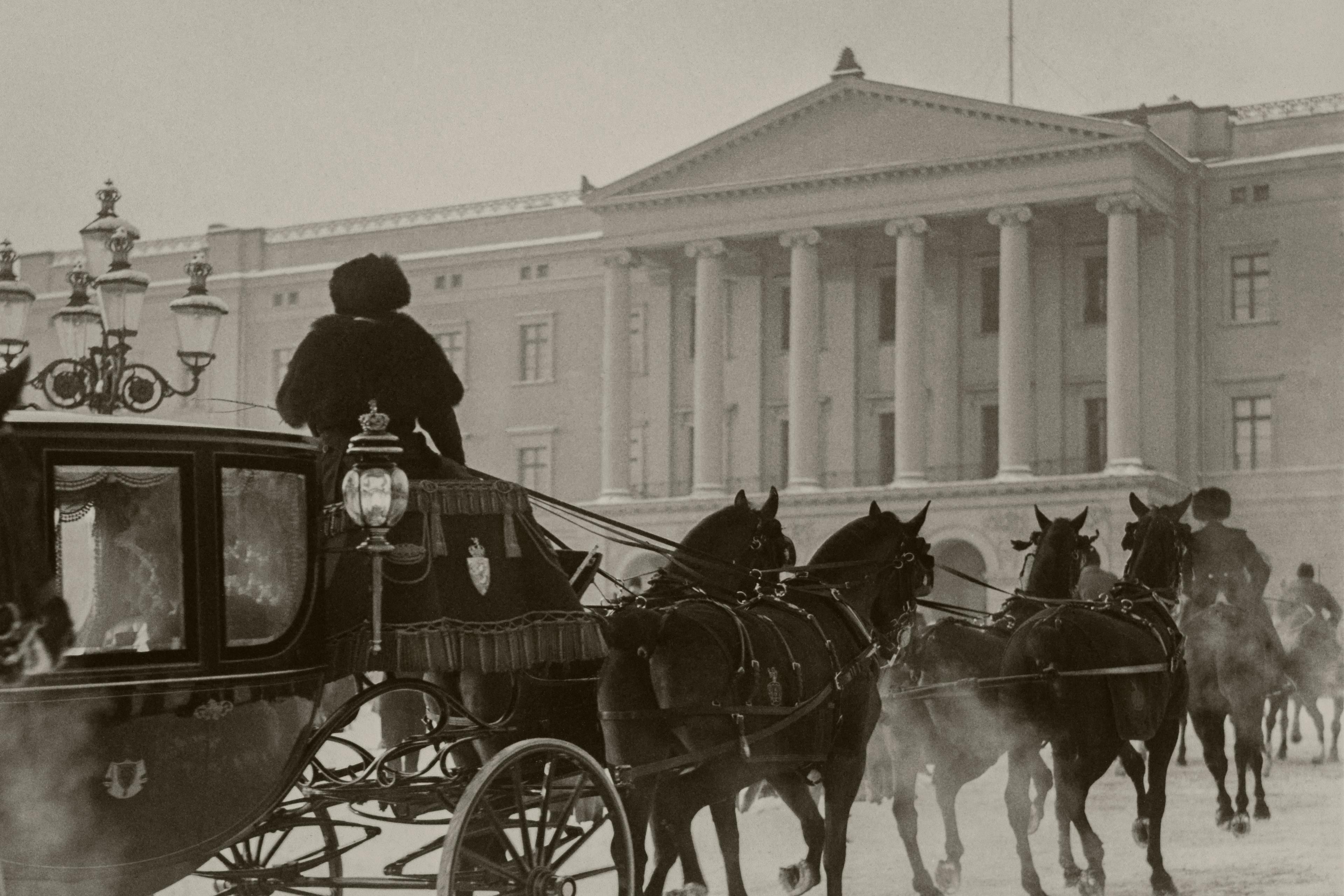Sort-hvittbilde av vognen trukket av fire mørke hester på vei opp den snølagte bakken inn på Slottsplassen. Slottets fasade i foran dem.