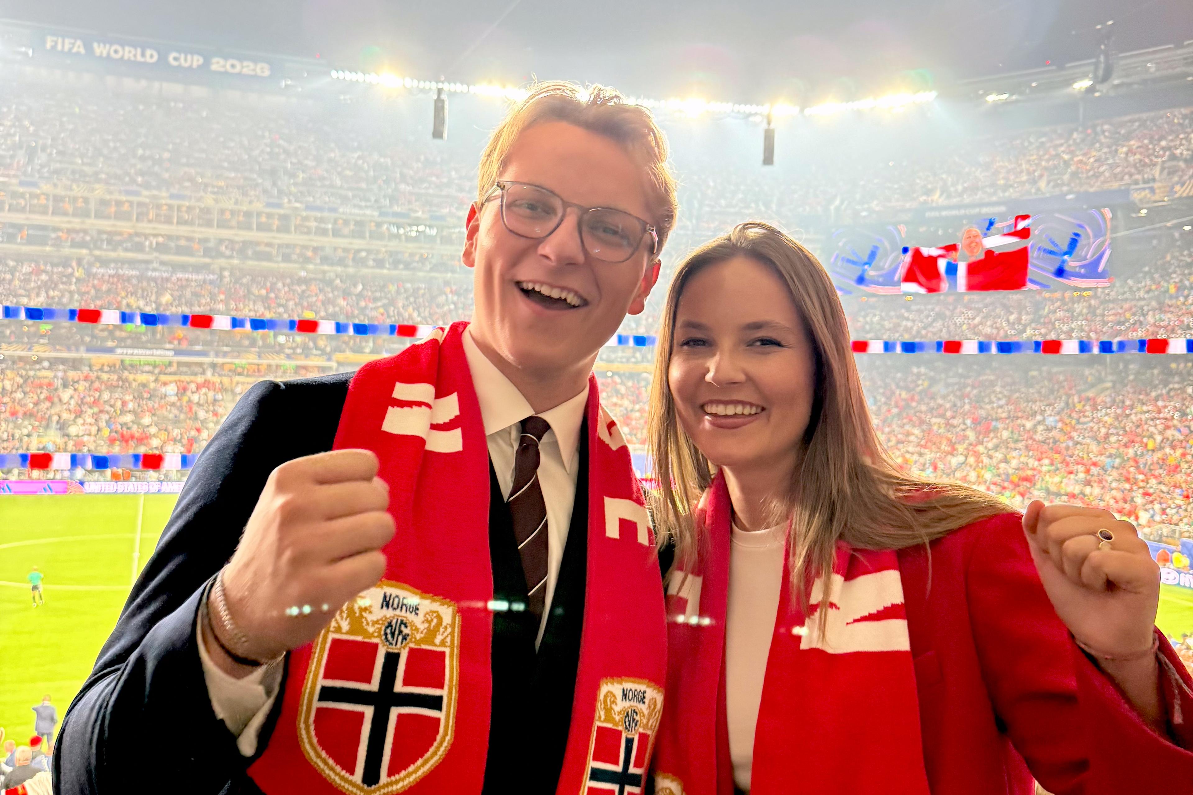 Prinsesse Ingrid Alexandra og Prins Sverre Magnus står sammen med norske fotballflagg rundt halsen. De smiler bredt og har en fotballstadion full av mennesker i rødt, hvitt og blått bak seg.