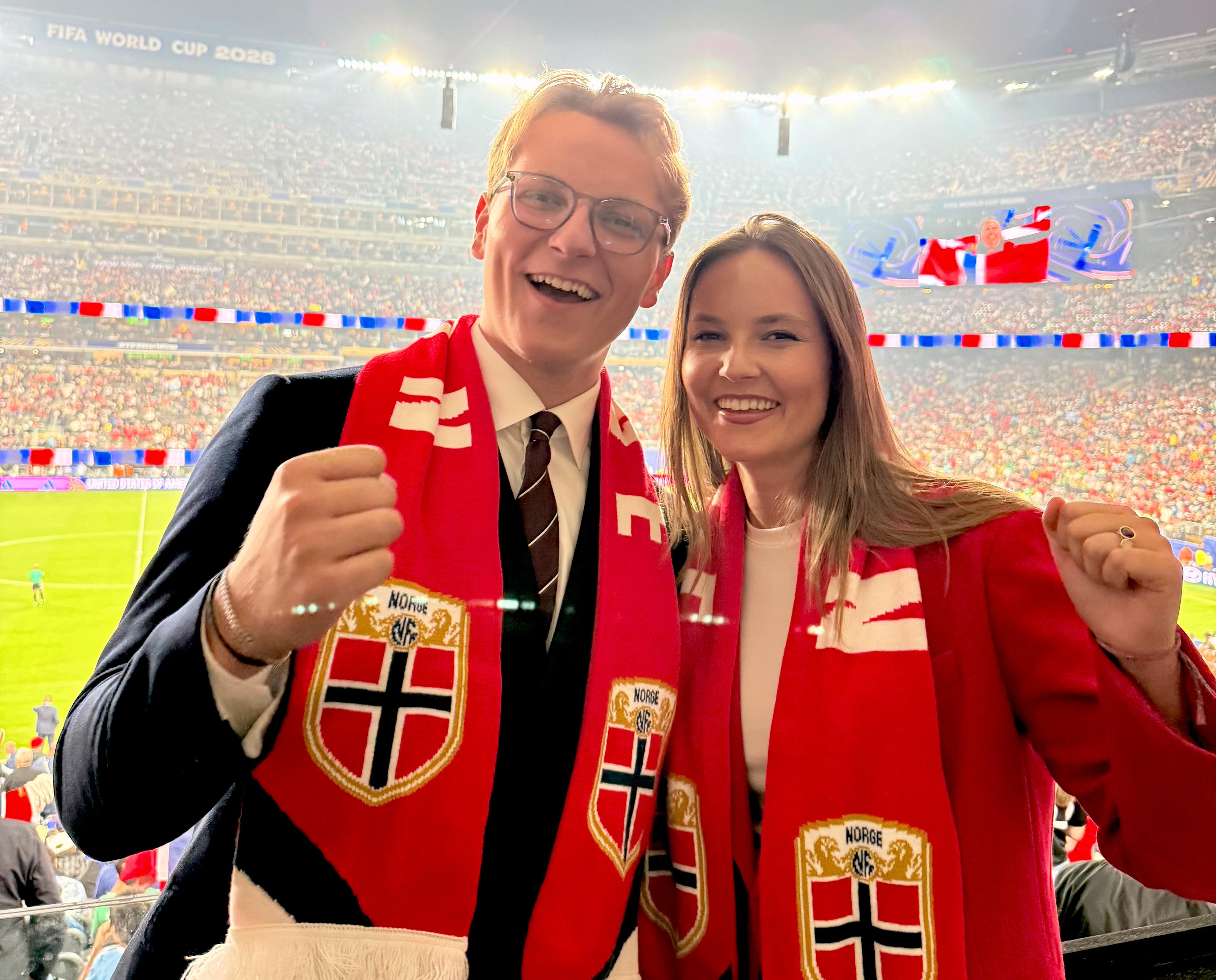 Prinsesse Ingrid Alexandra og Prins Sverre Magnus står sammen med norske fotballflagg rundt halsen. De smiler bredt og har en fotballstadion full av mennesker i rødt, hvitt og blått bak seg.