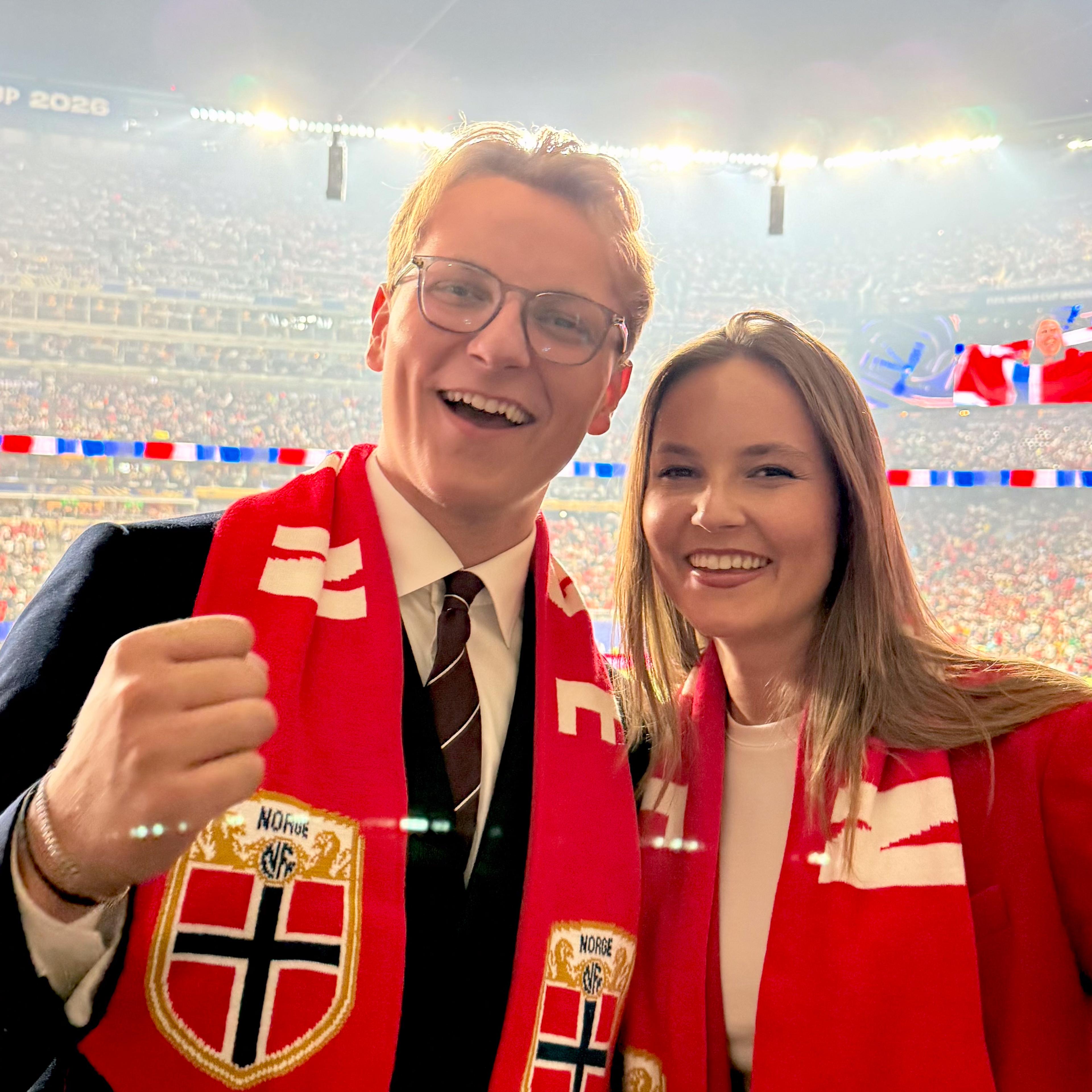Prinsesse Ingrid Alexandra og Prins Sverre Magnus står sammen med norske fotballflagg rundt halsen. De smiler bredt og har en fotballstadion full av mennesker i rødt, hvitt og blått bak seg.