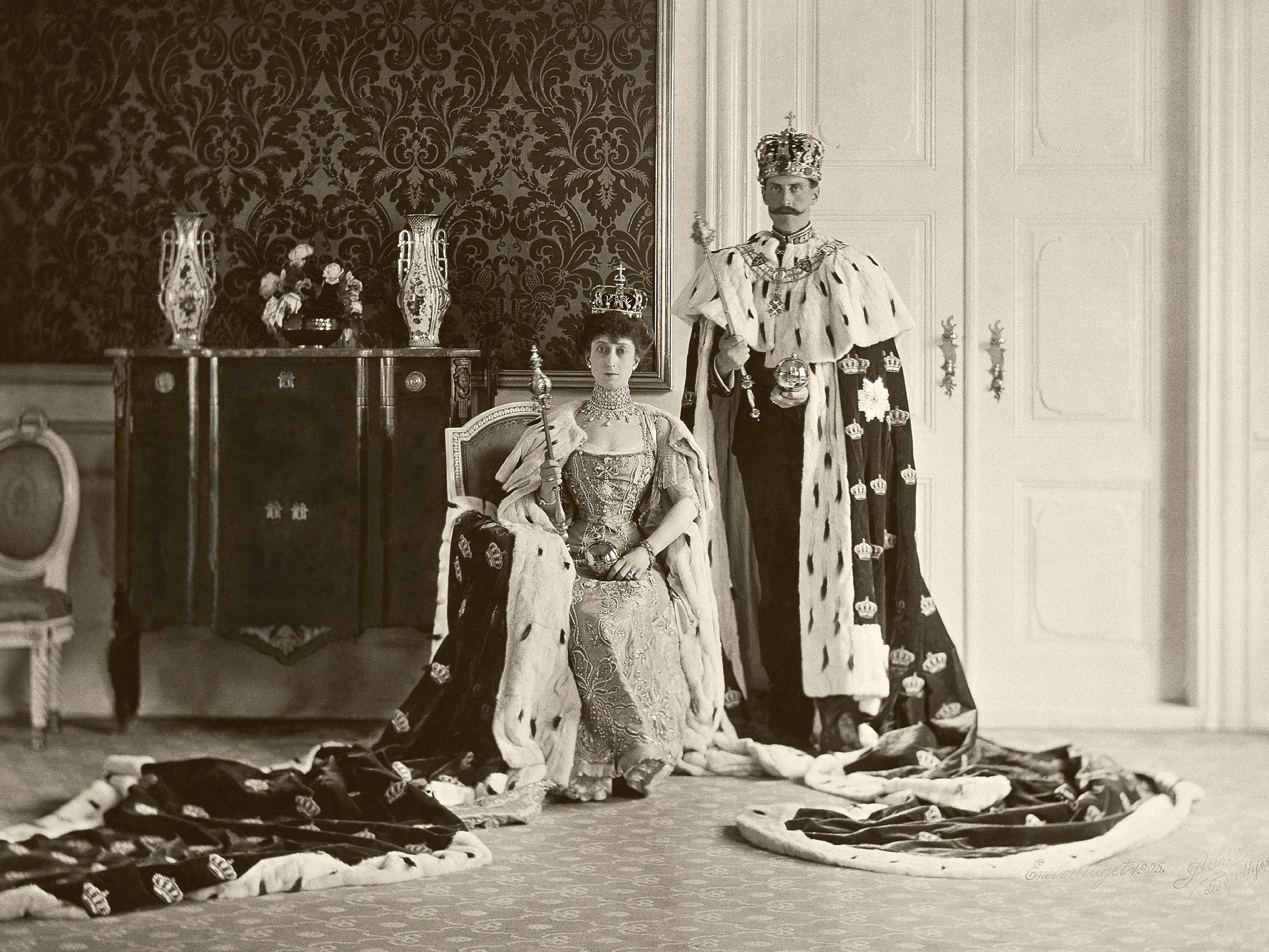 Sort-hvitt bilde av Kongeparet med kroner på hodet og kroningskapper i hermelin. Dronningen sitter på en stol mens Kongen står ved siden av henne. Begge holder et septer i hendene.