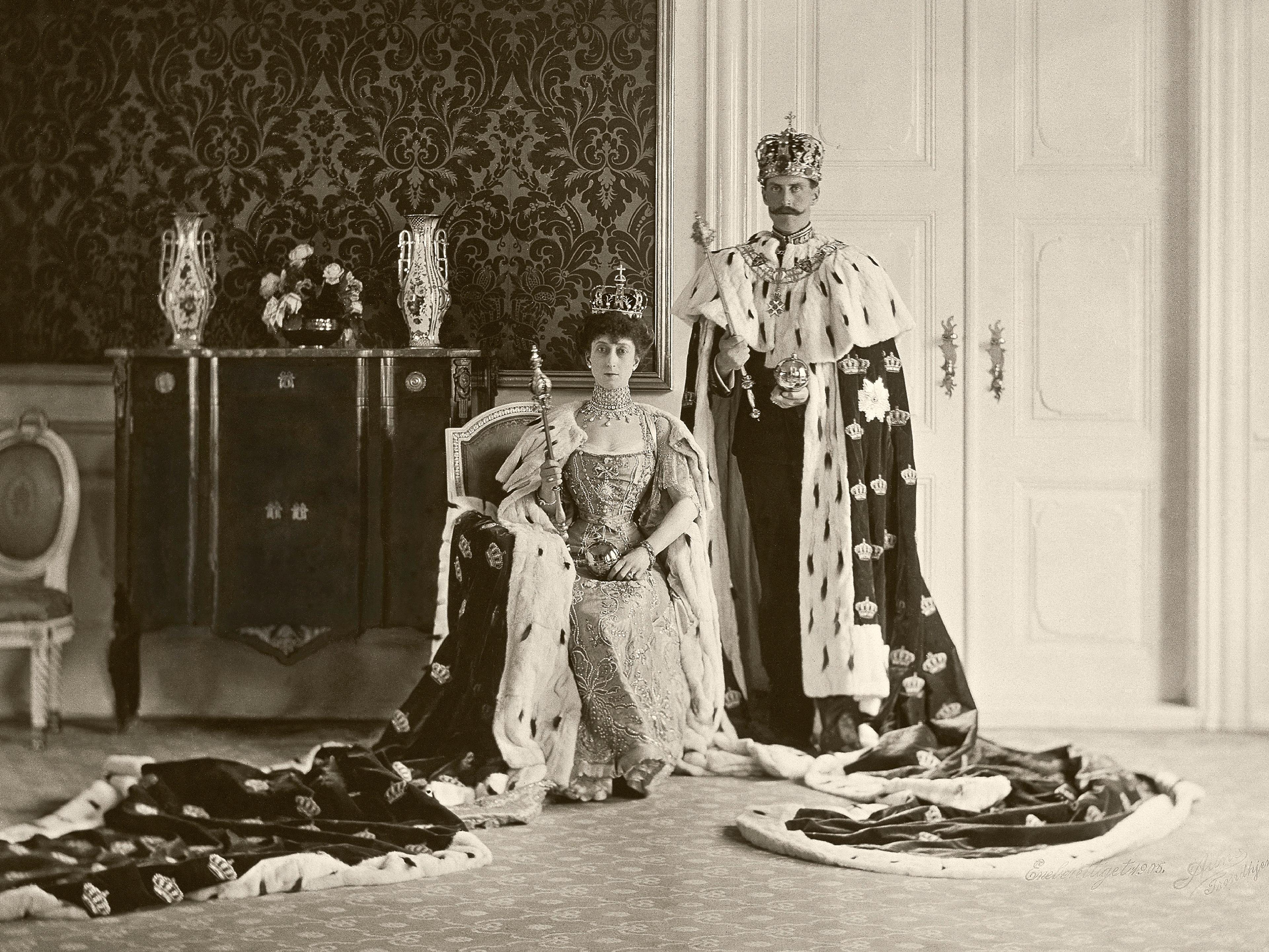 Sort-hvitt bilde av Kongeparet med kroner på hodet og kroningskapper i hermelin. Dronningen sitter på en stol mens Kongen står ved siden av henne. Begge holder et septer i hendene.