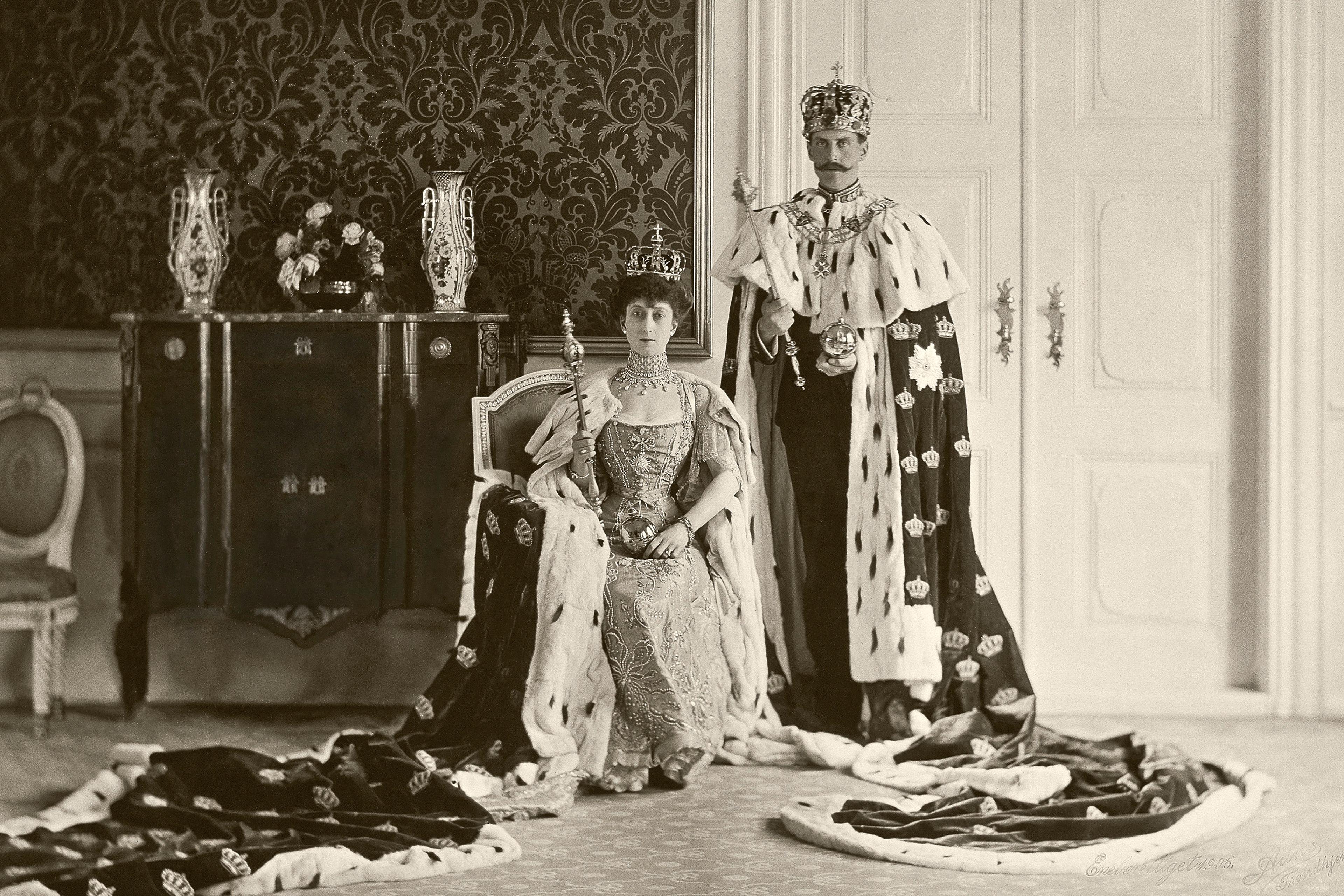 Sort-hvitt bilde av Kongeparet med kroner på hodet og kroningskapper i hermelin. Dronningen sitter på en stol mens Kongen står ved siden av henne. Begge holder et septer i hendene.