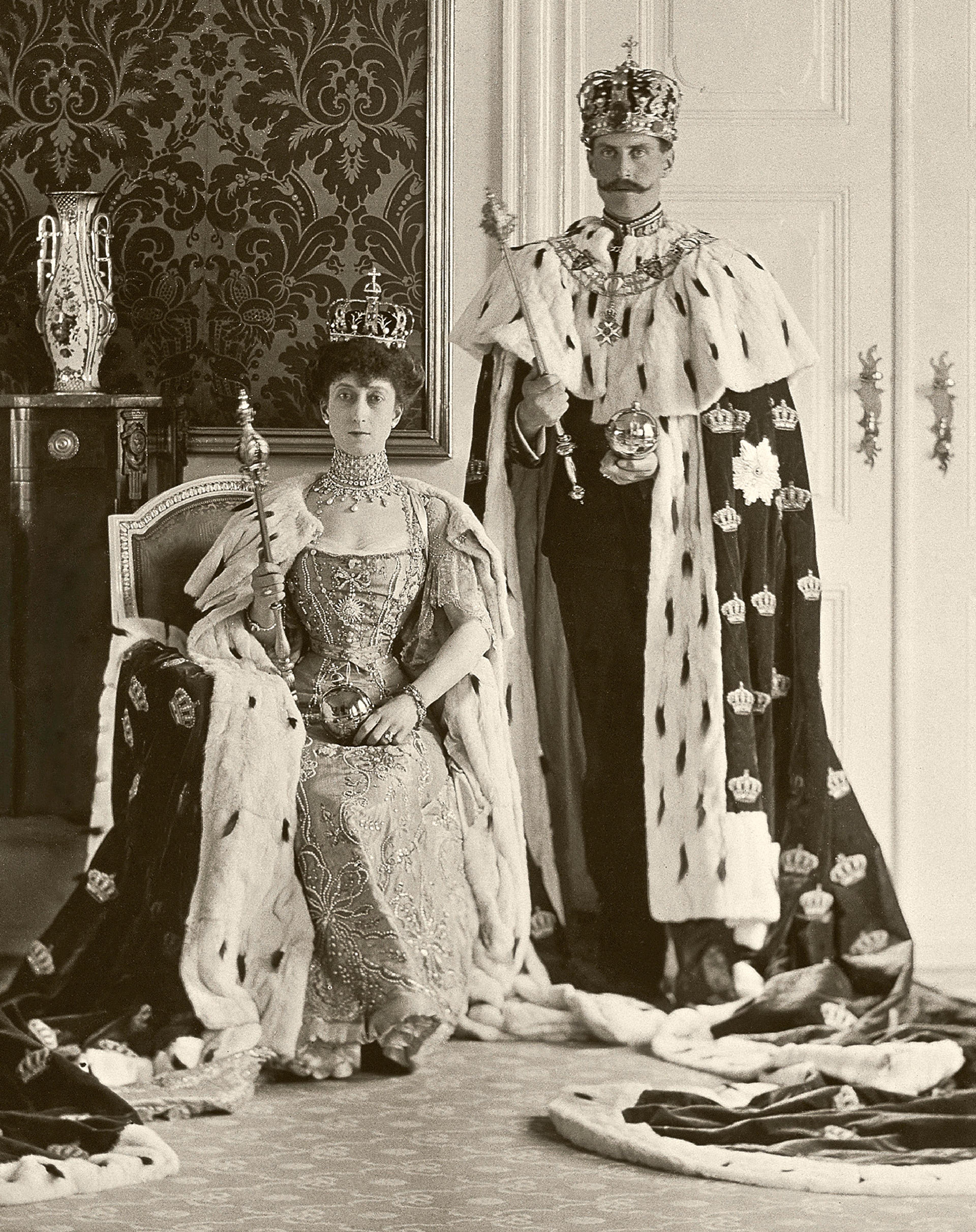 Sort-hvitt bilde av Kongeparet med kroner på hodet og kroningskapper i hermelin. Dronningen sitter på en stol mens Kongen står ved siden av henne. Begge holder et septer i hendene.