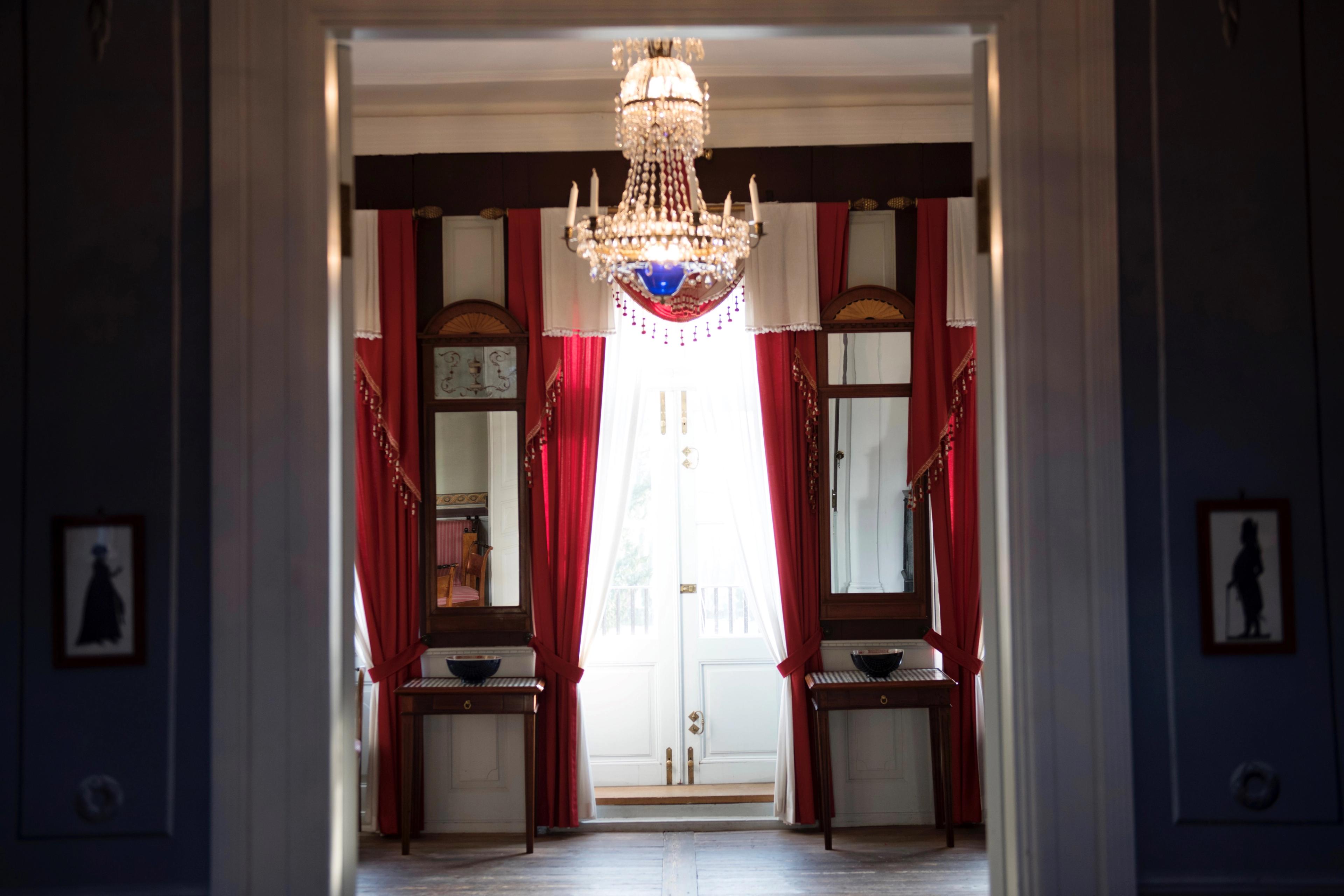 Gjennom en åpen dør ser vi inn i et rom med røde draperier og lysekrone