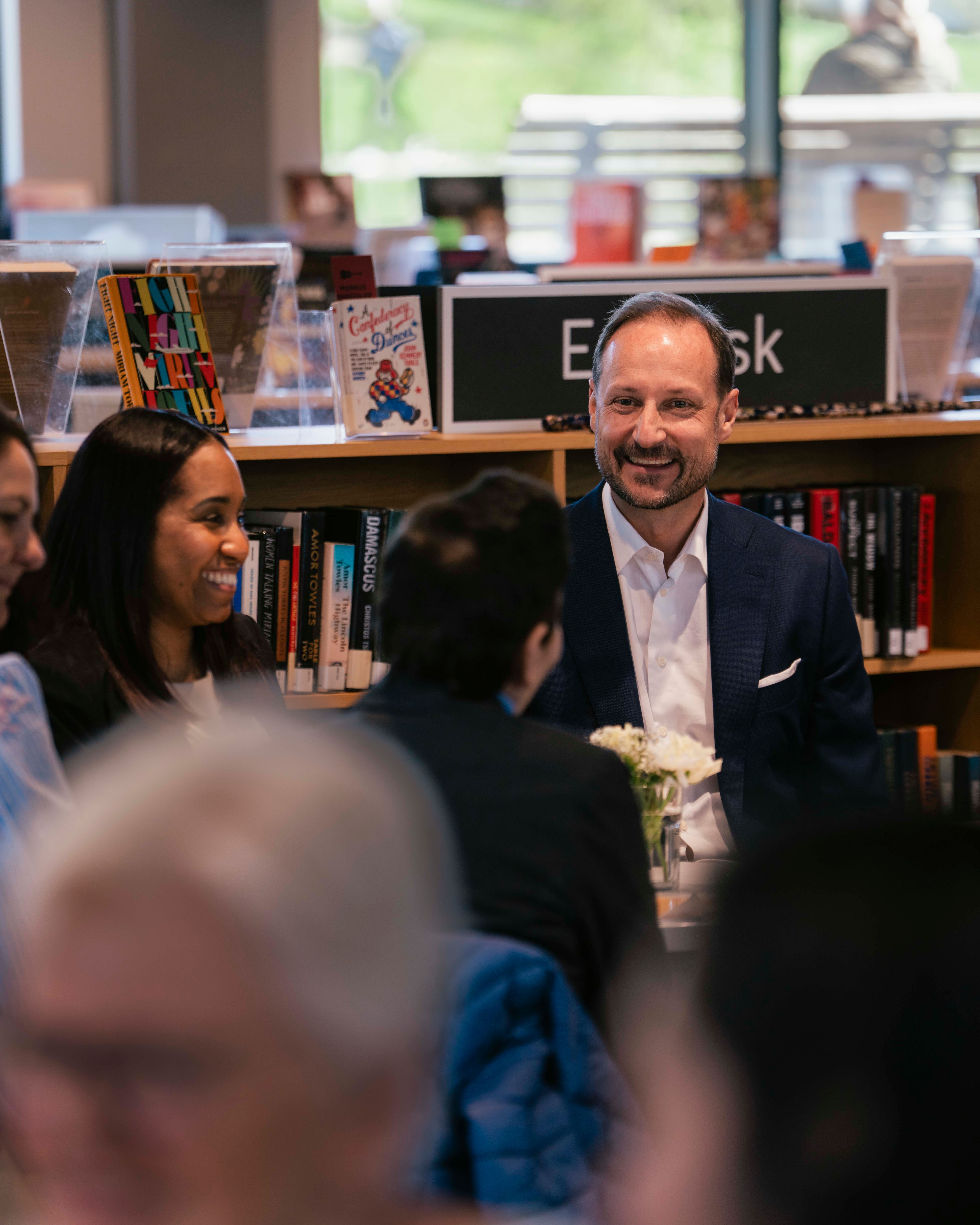 Kronprins Haakon sitter i en gruppe med mennesker rundt et bord på inne på et bibliotek, med bøker i bakgrunnen.