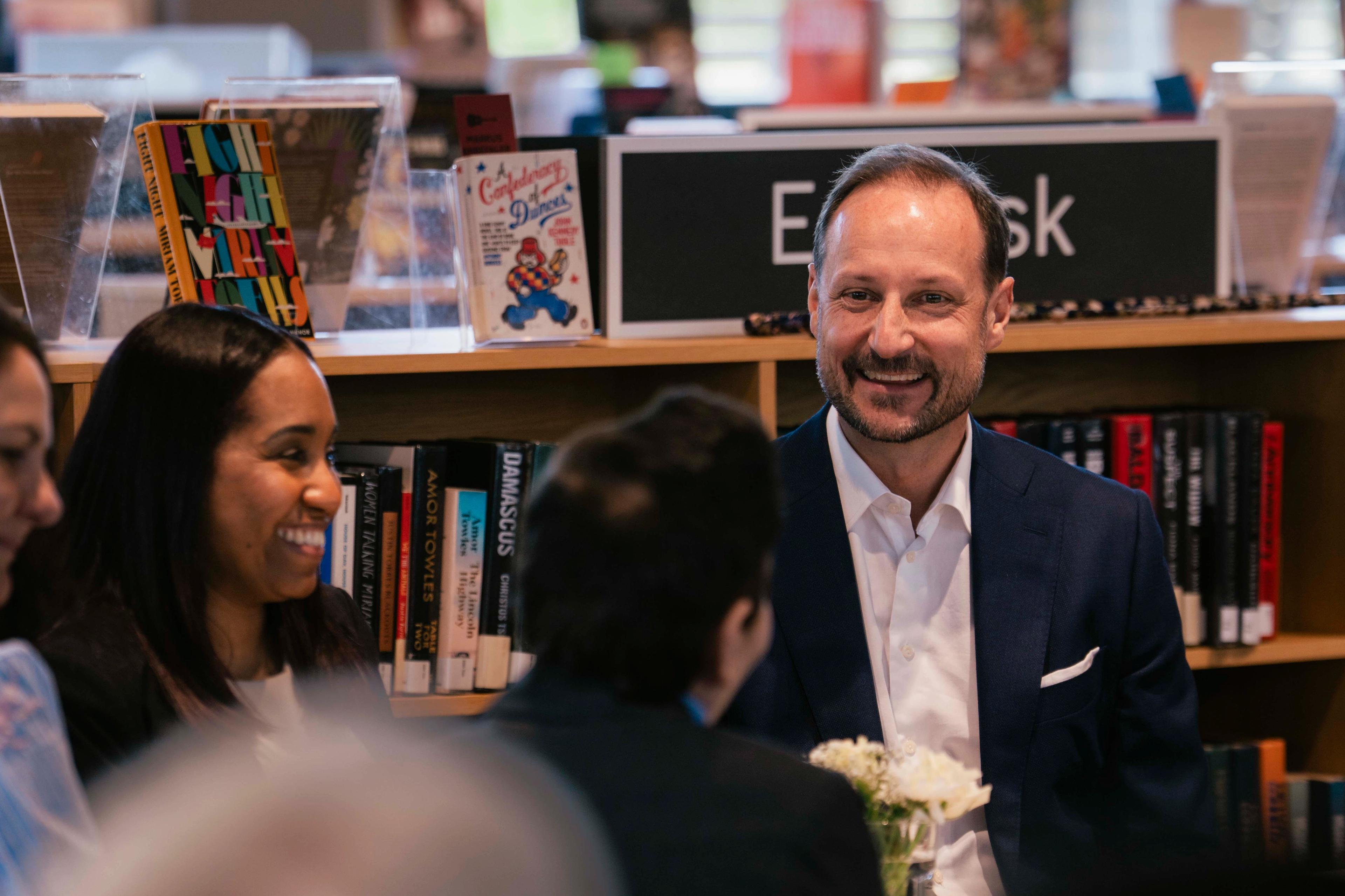 Kronprins Haakon sitter i en gruppe med mennesker rundt et bord på inne på et bibliotek, med bøker i bakgrunnen.