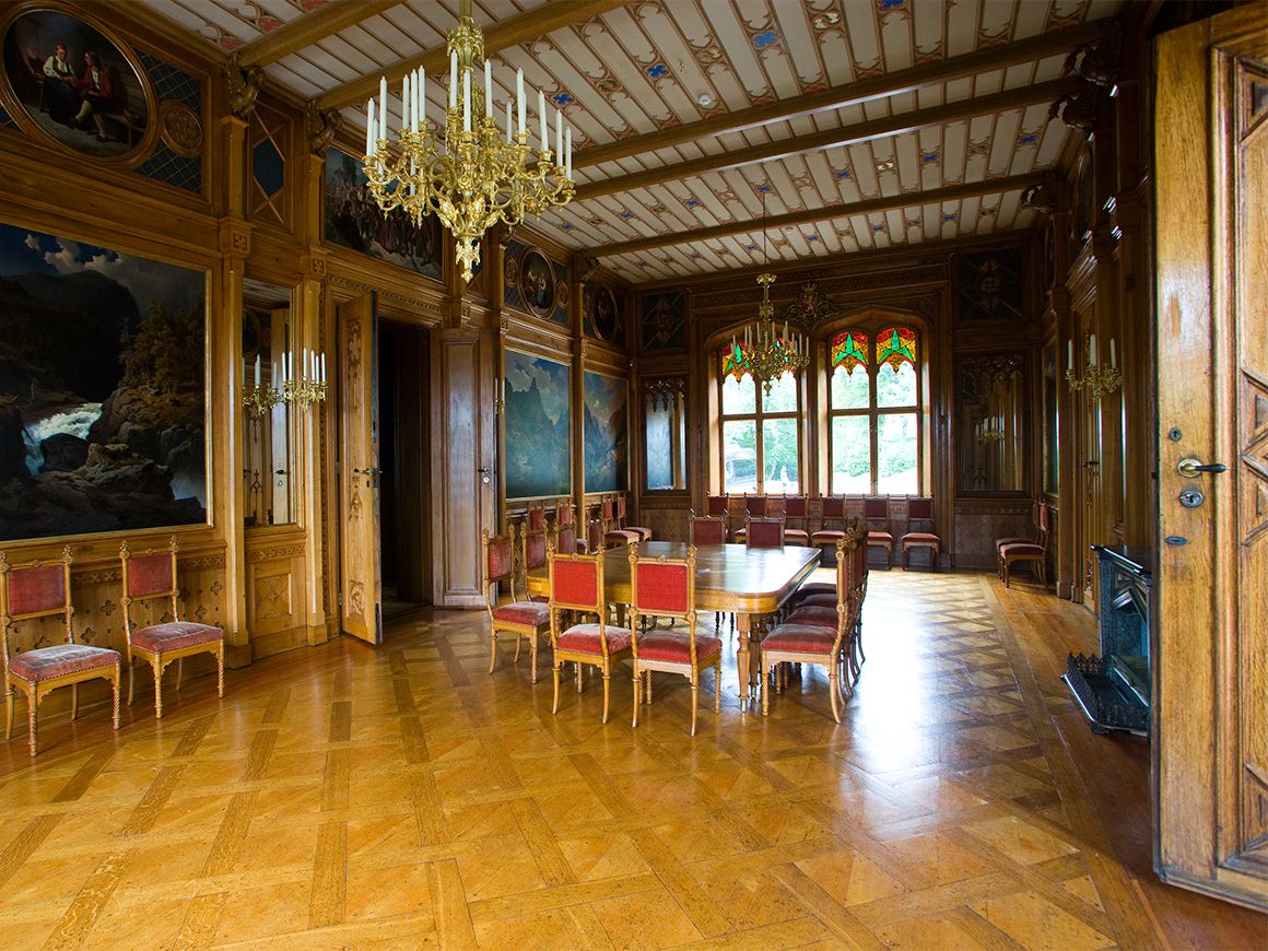 Bord og stoler står midt i et rom med store vinduer og vegger utsmykket med malerier.